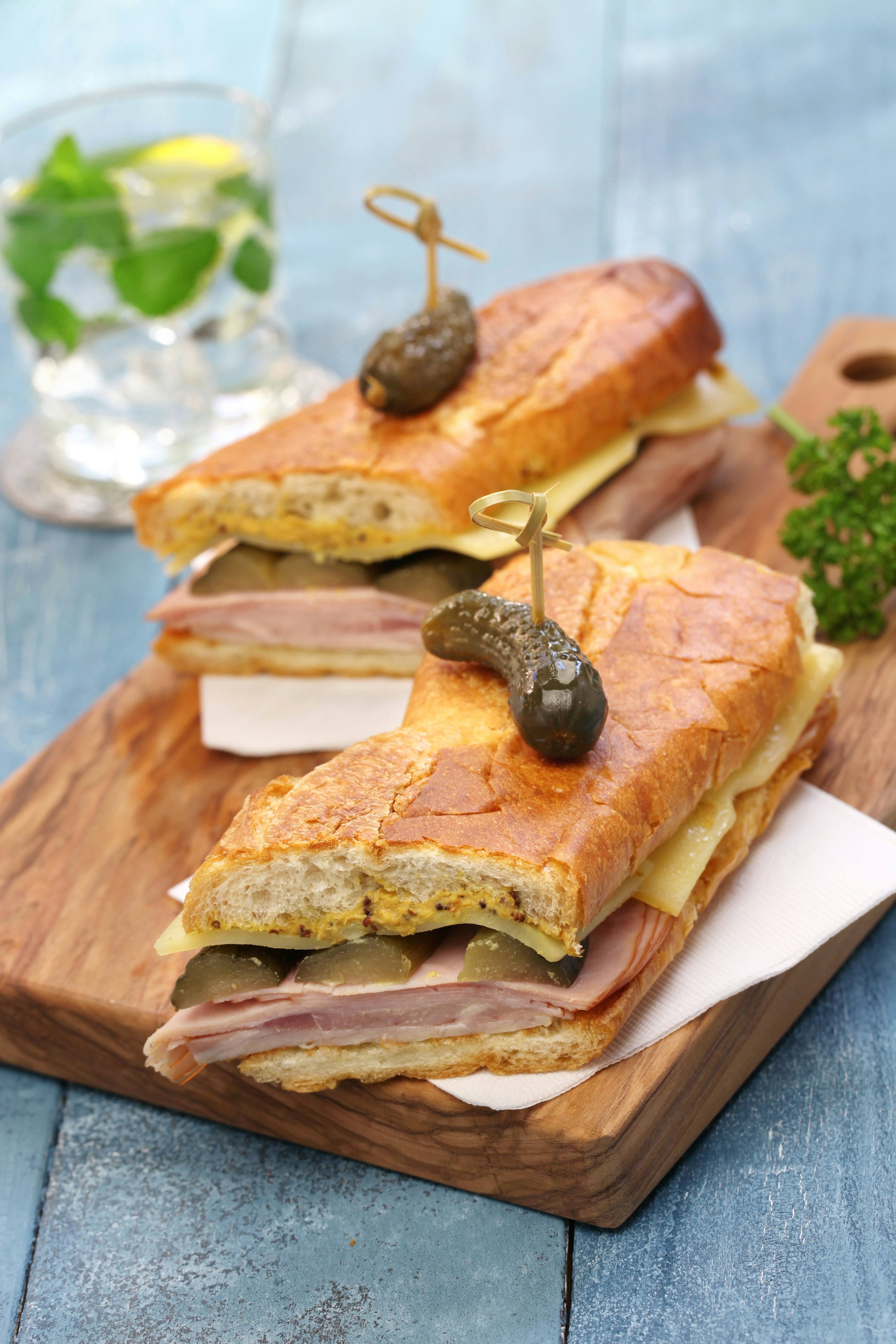 A long pressed sandwich is cut in half and arranged artfully on a chopping board. The sandwich is stuffed with ham, cheese, pork and pickles. A pickle is skewered through each half of the sandwich a wooden stick.