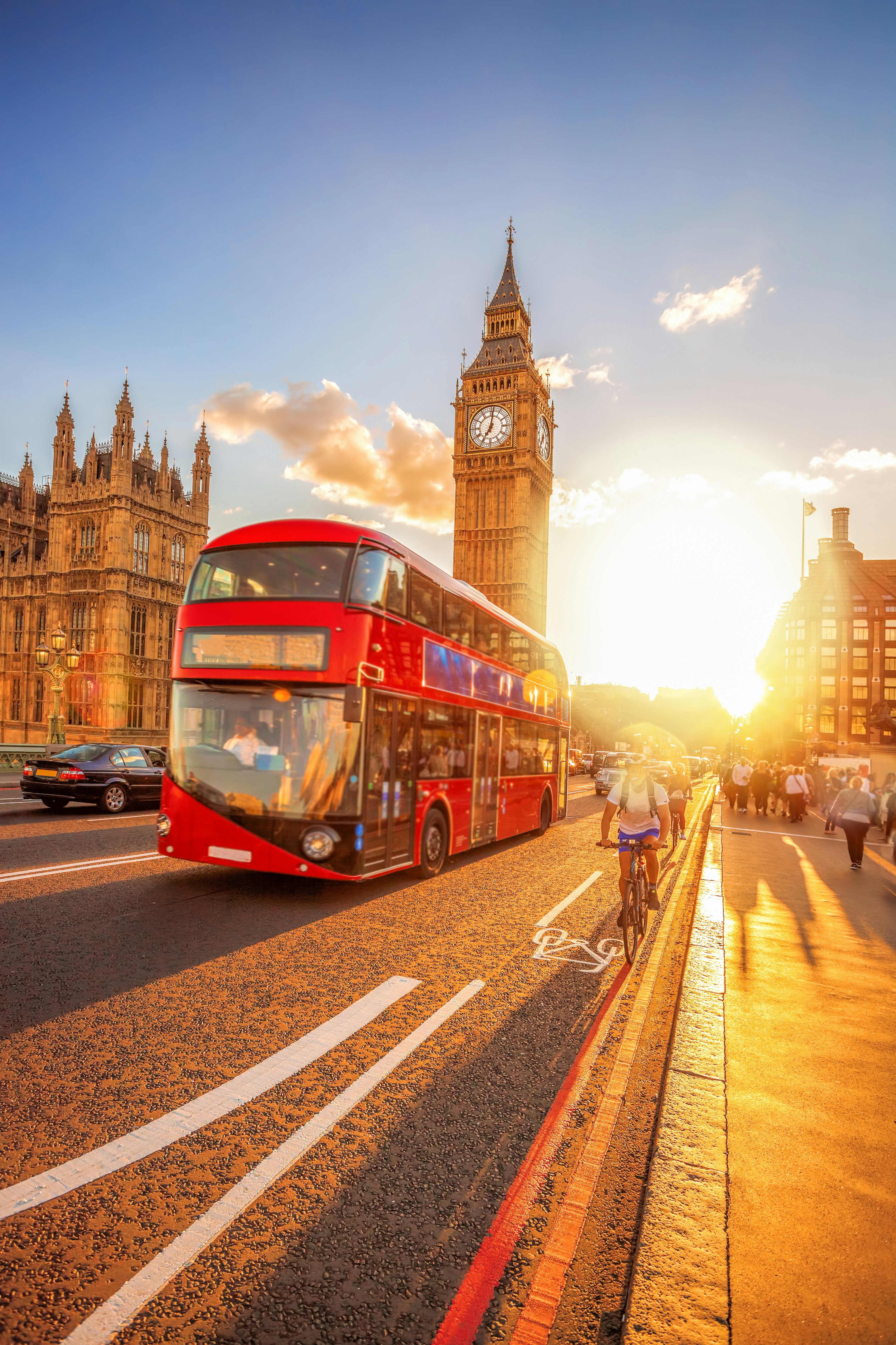 A golden-hour photo taken on Westminster Bridge in London. The sun is low in the sky, casting a glow over the scene of Big Ben, the Houses of Parliament, a red London bus, and people walking and cycling over the bridge.