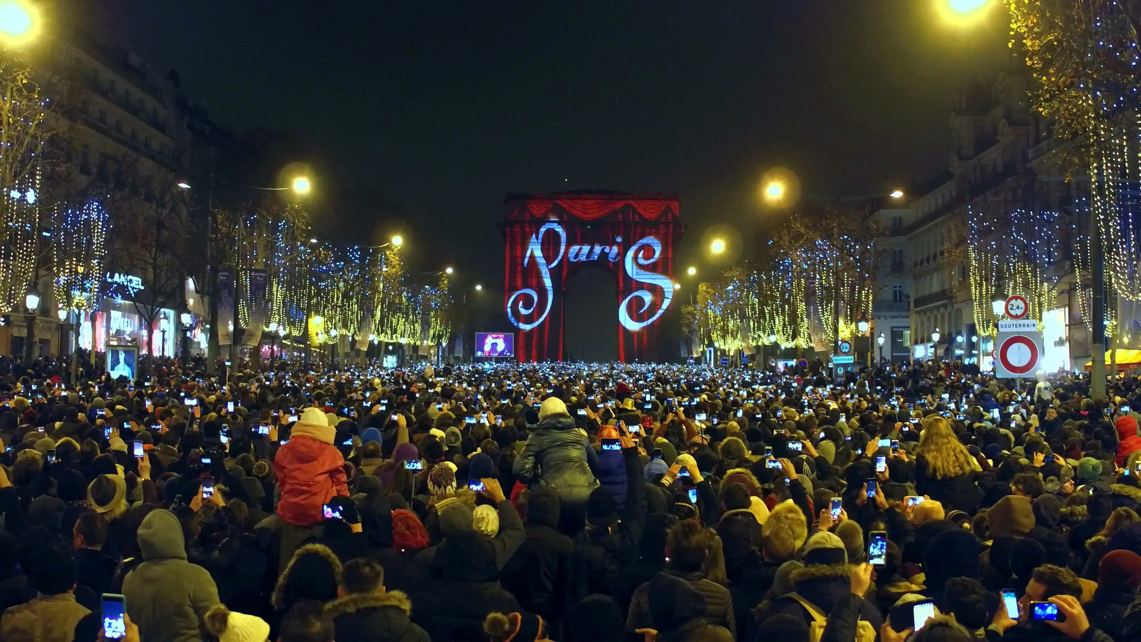 En skara av tusentals samlas på natten på Champs-Élysées i Paris, vänd mot Triumfbågen, som är upplyst för att likna en teaterridå med 