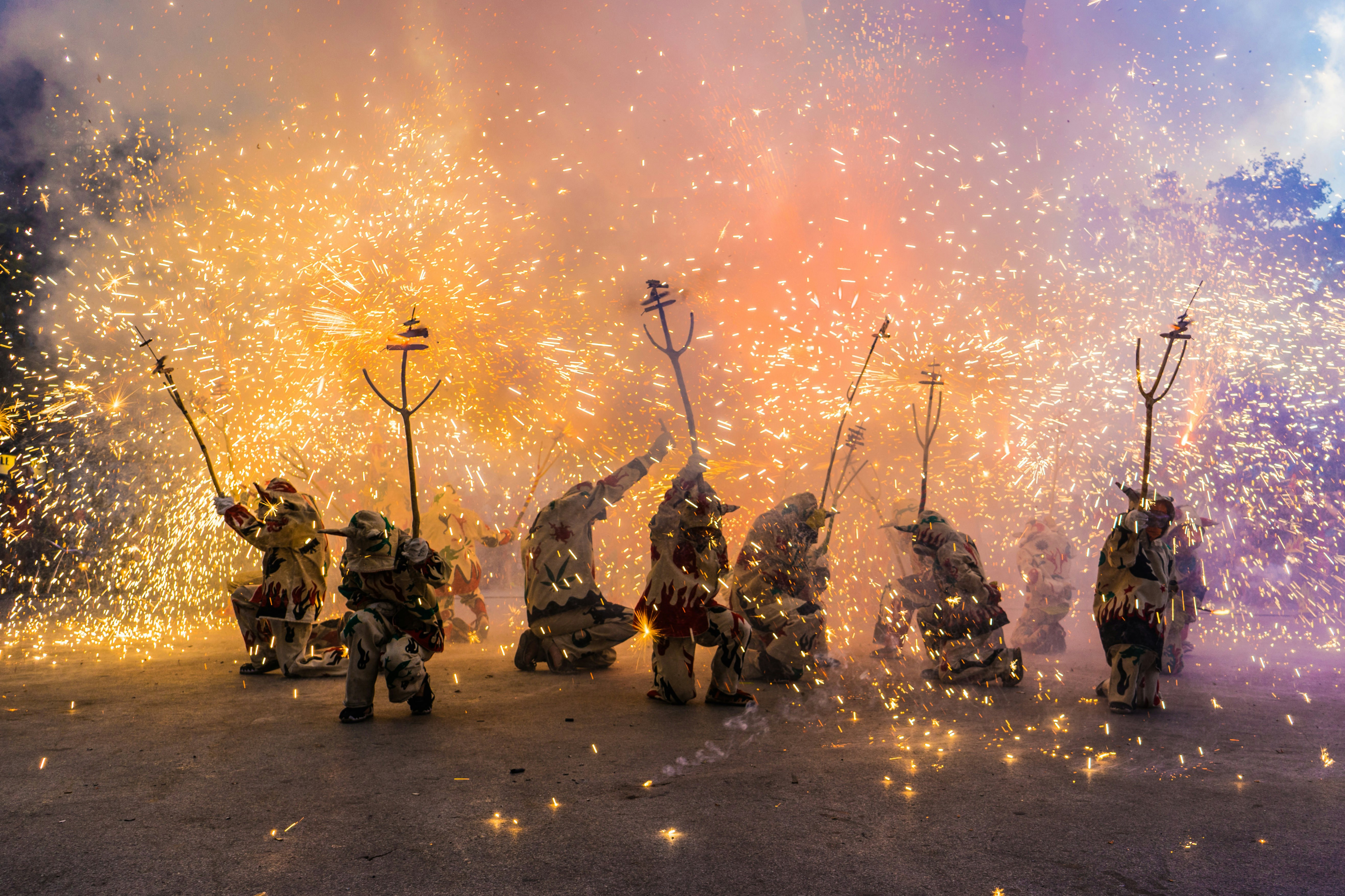 People dressed as devils kneel on the floor brandishing pitchforks while fireworks erupt around them.