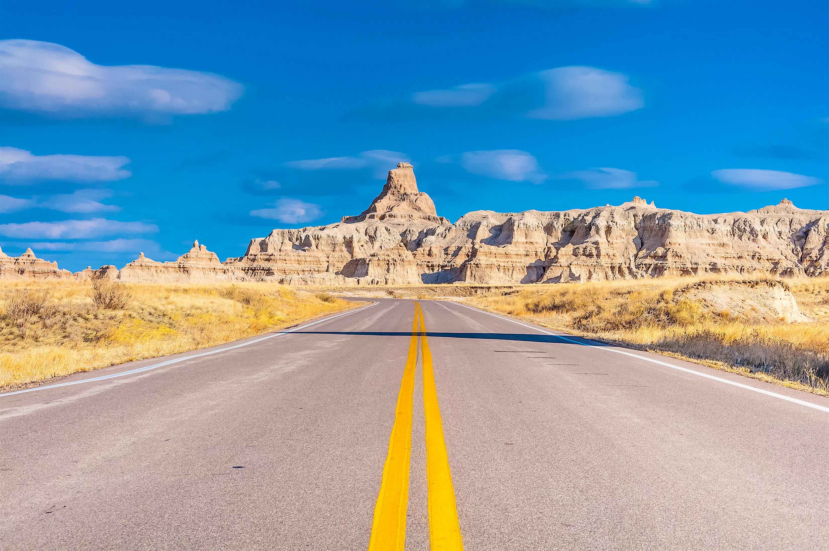 shutterstockRF_560780143.jpg A straight empty road stretches through Badlands National Park, with large rock formations in the distance against a blue sky