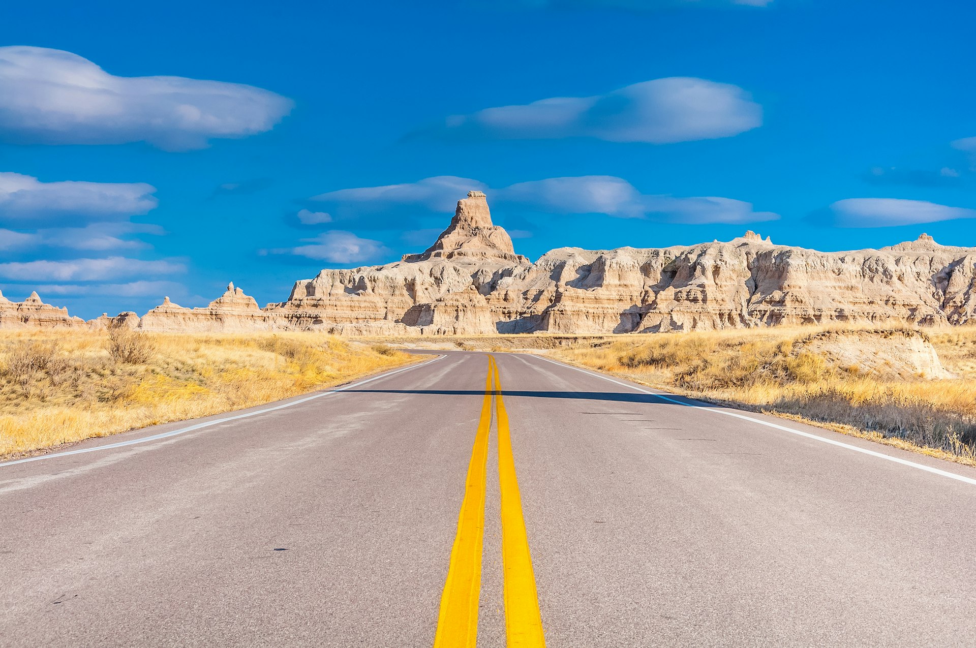 A straight empty road stretches through Badlands National Park, with large rock formations in the distance against a blue sky