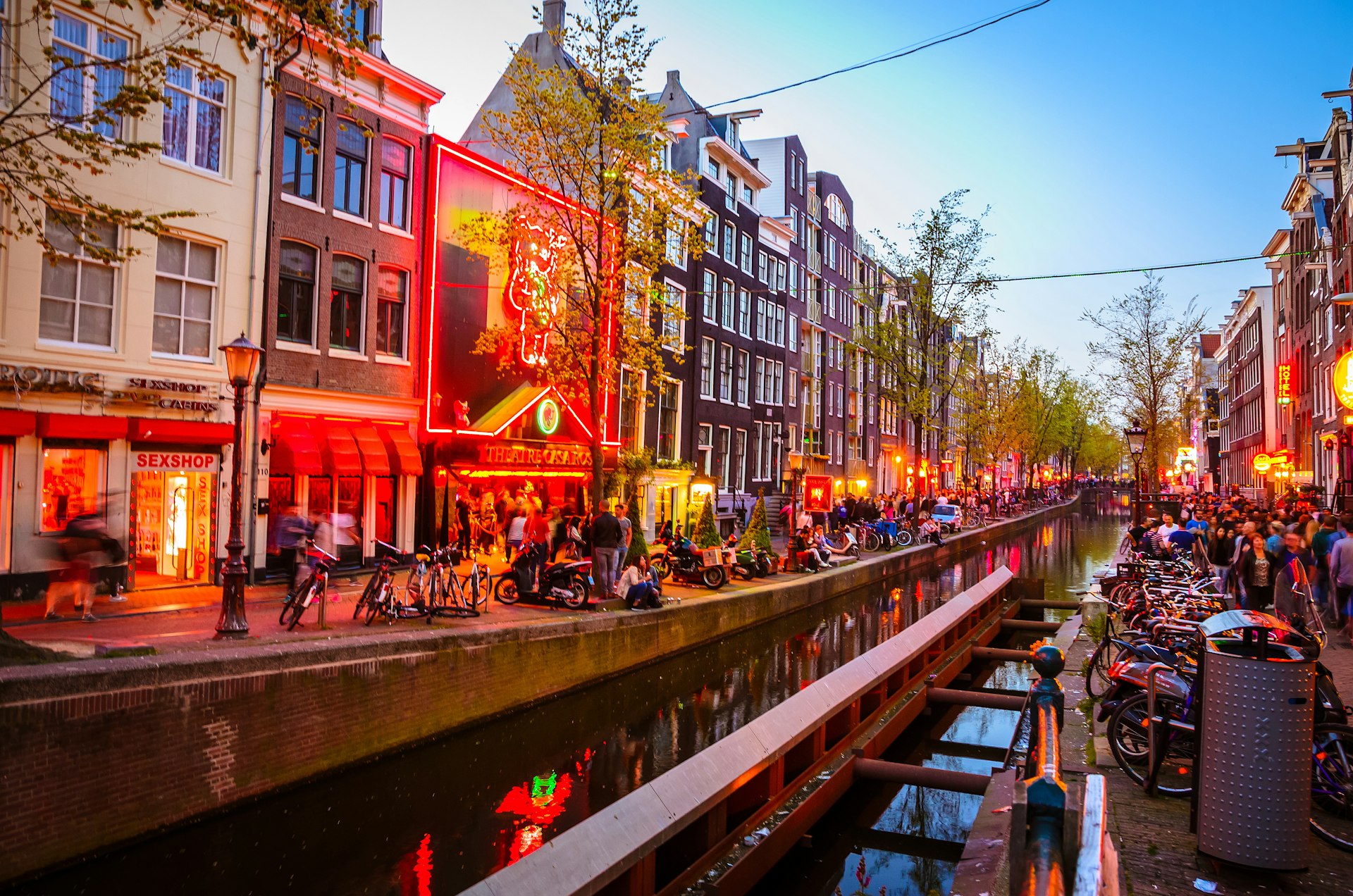 A canal cuts through the centre of Amsterdam's Red Light District, with bars and restaurants (lit in the iconic red lights) flanking the streets either side, while the roads are busy with people. 