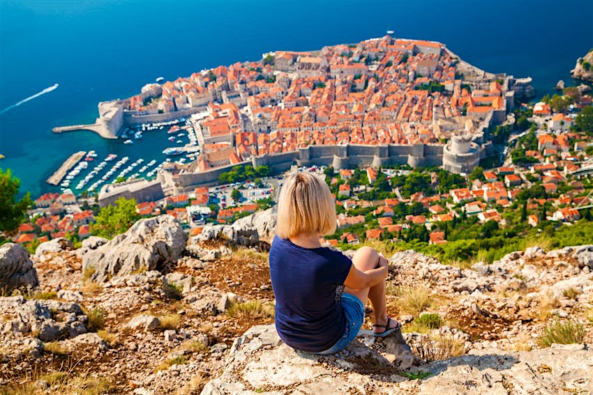 A girl sits at the top of a steep, rocky mountain, gazing down at the orange roofs of Dubrovnik far below, encircled by stone city walls with azure sea beyond.