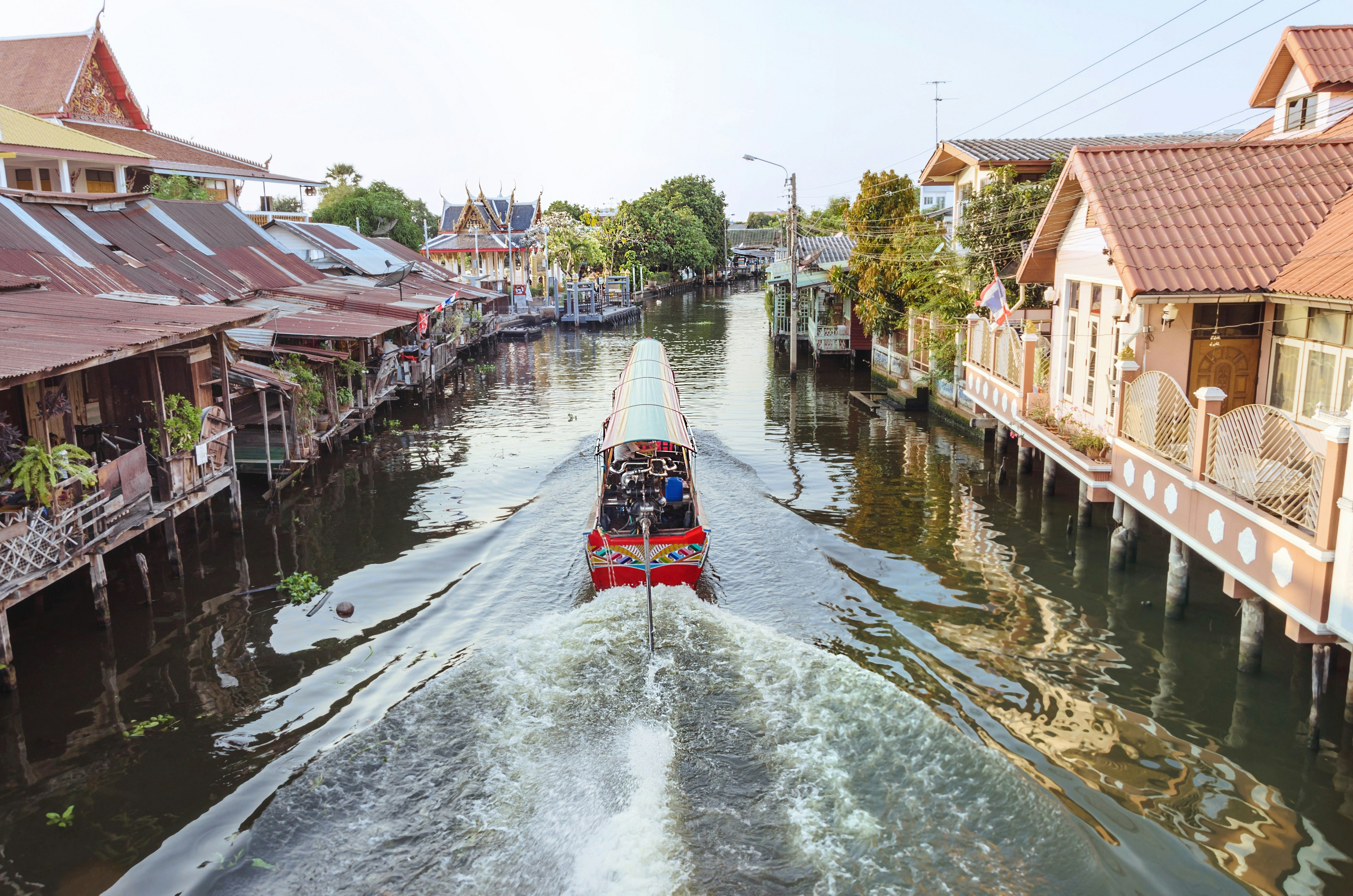 A boat cruises through the centre of a canal in Bangkok. The narrow canal is flanked by wooden houses on stilts above the waterline.