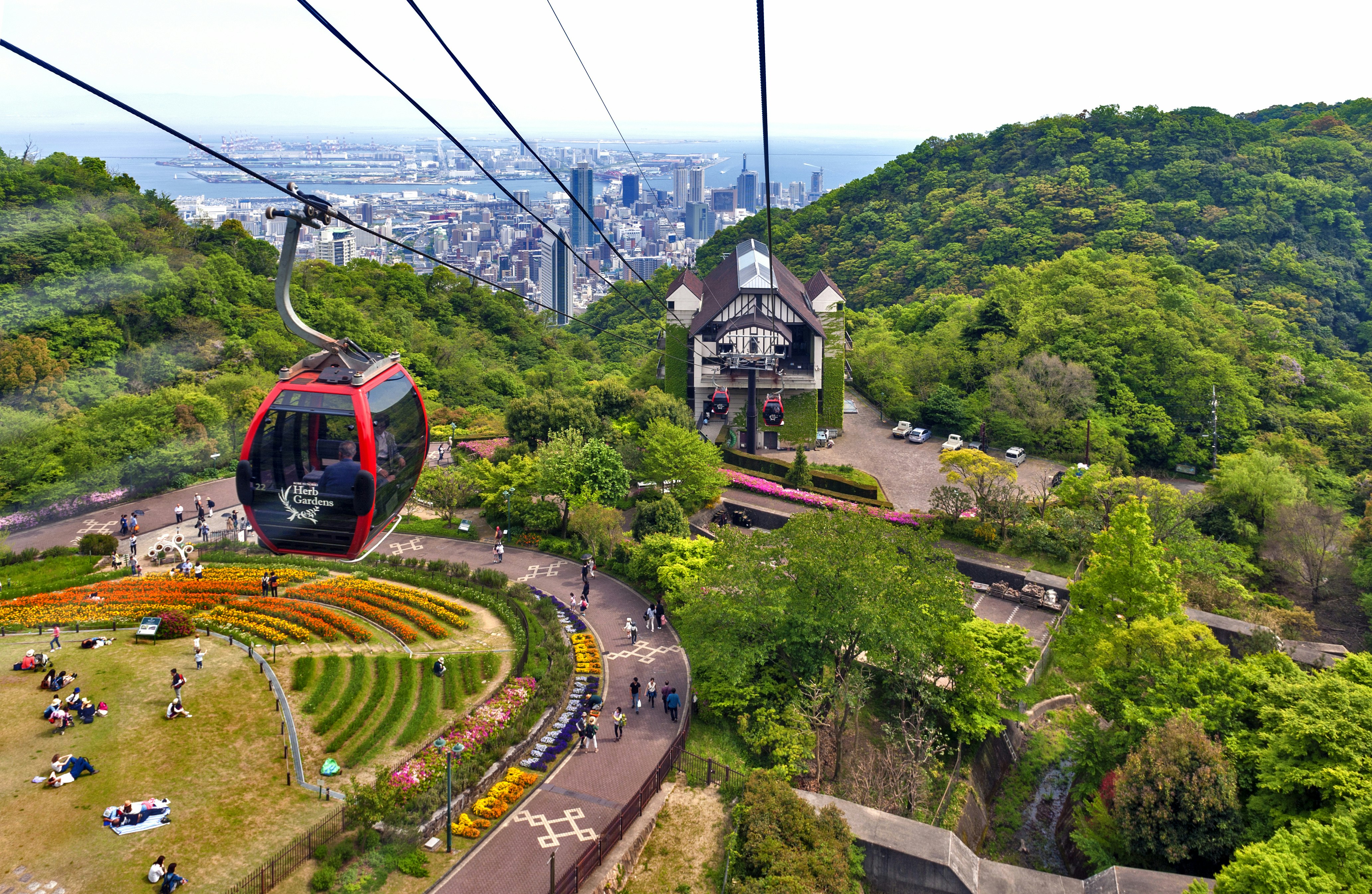 A red cable car carriage travels along a wire near the city of Kōbe. Below people sit beside a green manicured garden and in the background the city skyline is visible.