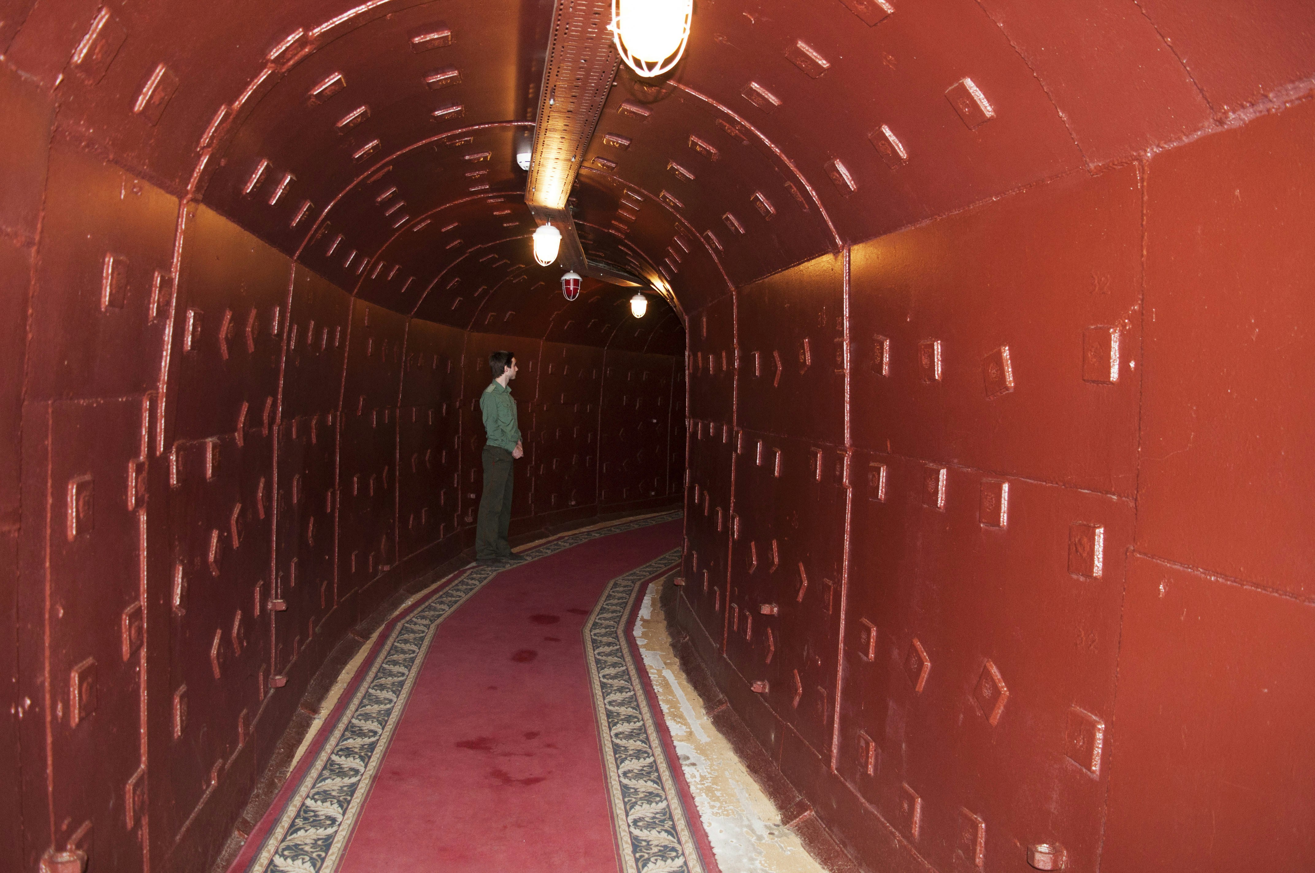 An underground tunnel lined with reddish-brown walls curves round a corner. A guide stands towards the bend.