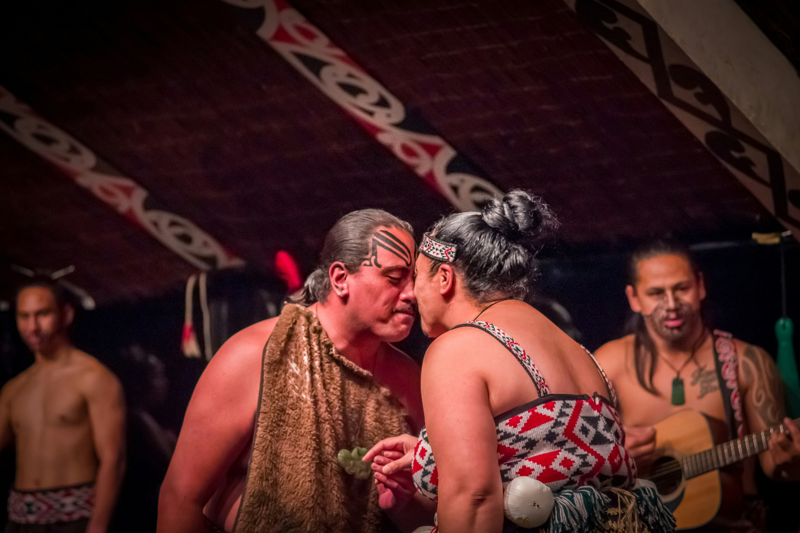 A Tamaki Māori couple is dancing. They are facing each other, holding hands and touching noses. They are wearing traditional clothing; the man has tan-coloured cloth draped over his torso, while the woman has a top with a red and grey geometric pattern. There is a Māori guitarist in the background.