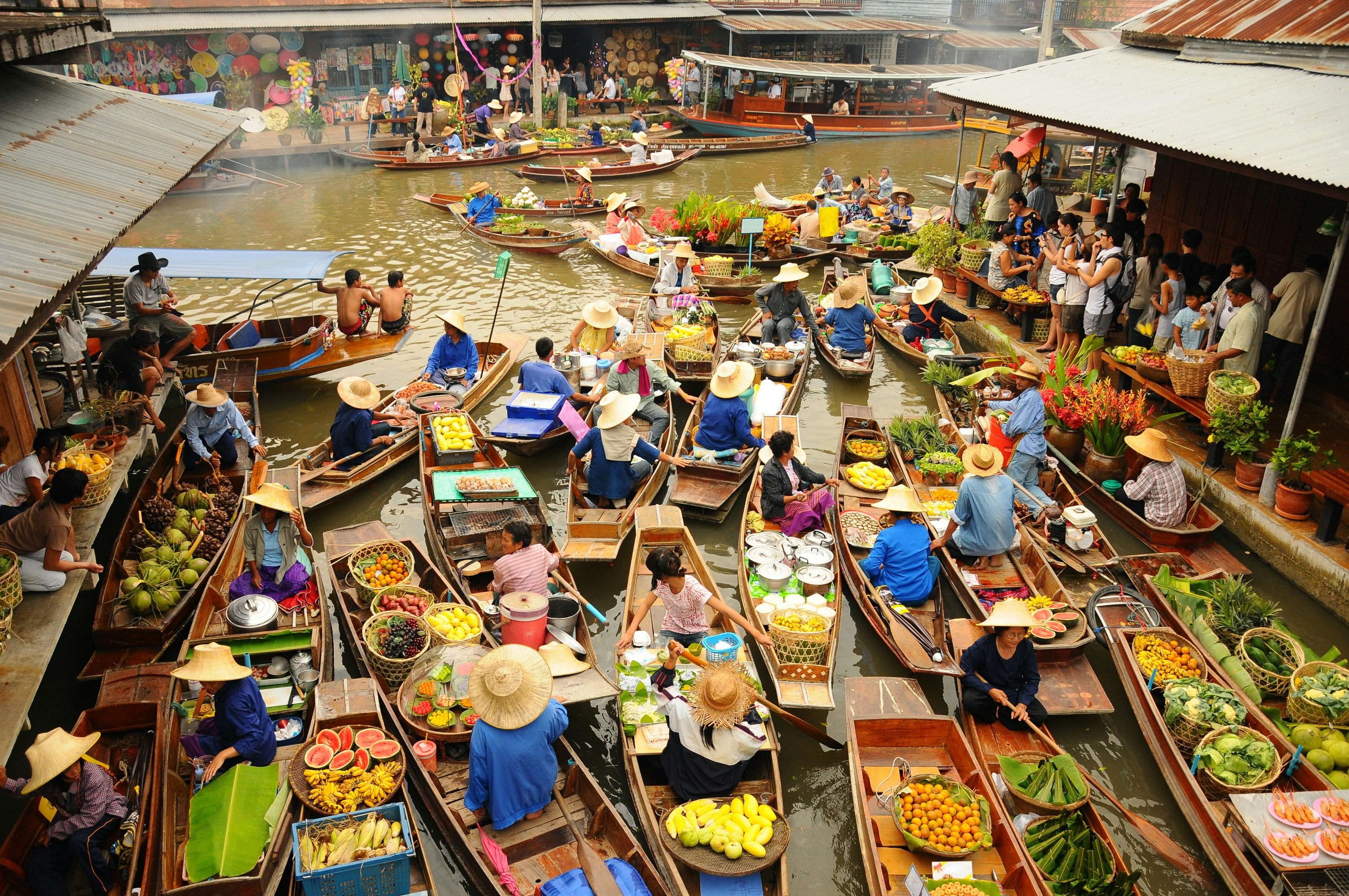 A vast amounts of small wooden boats gather to trade goods at  Amphawa floating market. Most of the boats sell food, specifically fruits. Tourist wielding cameras watch on. 