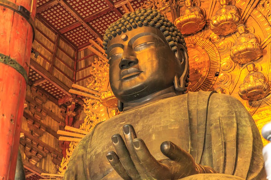 A huge bronze Buddha at the Todai-ji Temple in Nara; behind it is a large ornate gold dial with smaller Buddhas on it; there is an orange pillar next to the Buddha and wooden walls and ceiling. 