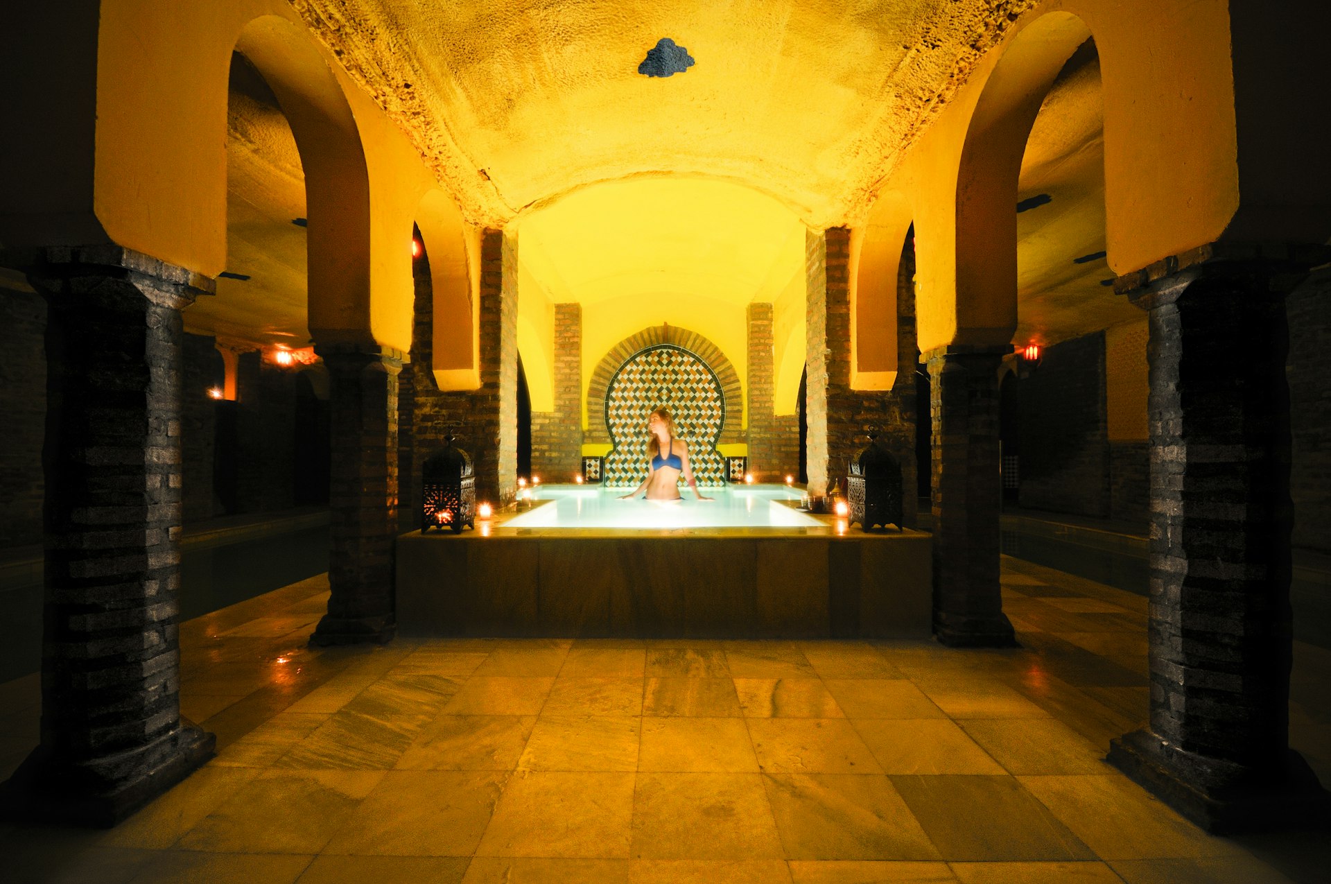 A woman is standing in a pool in a luxurious, romantically lit bathhouse. There are huge stone columns leading to the pool, and intricate tiles behind it.