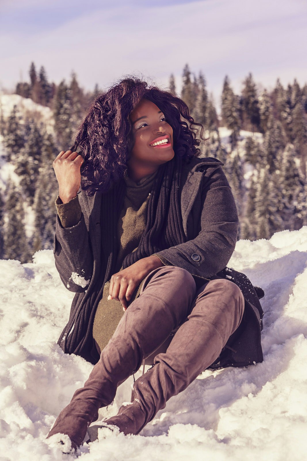 A woman sitting in crisp white snow in front of some snow-covered coniferous trees. She is playing with her hair and looking off to the side, smiling. She is dressed stylishly in a wool dress, warm coat, scarf, and over-the-knee boots.