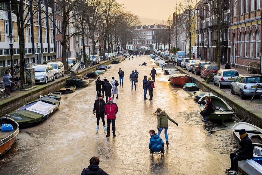 Människor som åker skridskor på en frusen Amsterdam-kanal;  kanalen kantas av båtar på båda sidor, och bredvid kanalen finns rader av bilar och radhus.  Amsterdam vinter
