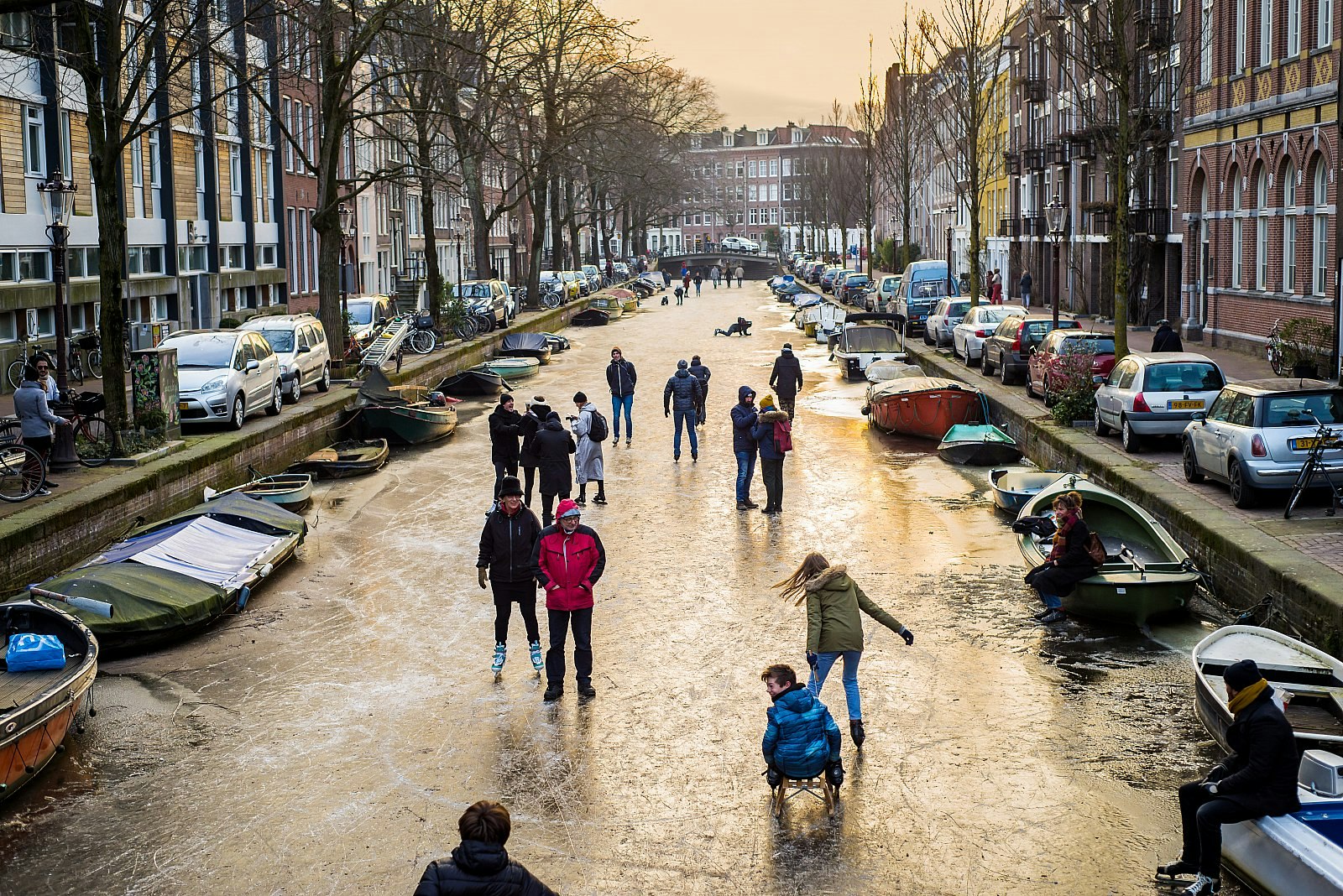 People ice-skating on a frozen Amsterdam canal; the canal is lined with boats on either side, and next to the canal are rows of cars and townhouses. Amsterdam winter