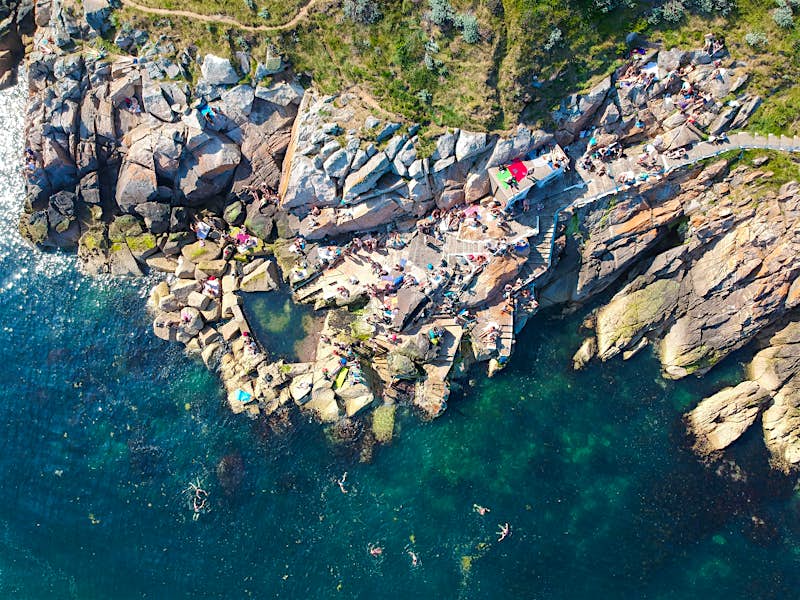 A birds-eye view of the Vico, an outdoor swimming spot near Dublin. Swimmers lounge on the staggered rocks that lead down to the dark blue sea. A small saltwater pool is also visible amongst the rocks. Some people can be seen swimming in the sea.