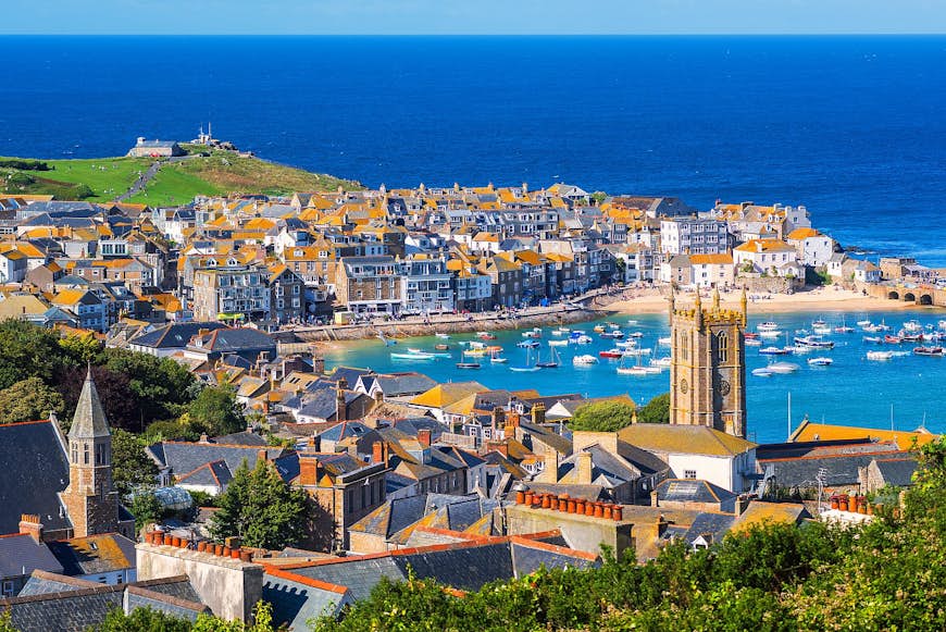 Looking over the stone buildings and slate rooftops of St Ives, which curves round a harbour onto a grassy headland, with the bright blue Atlantic Ocean beyond.