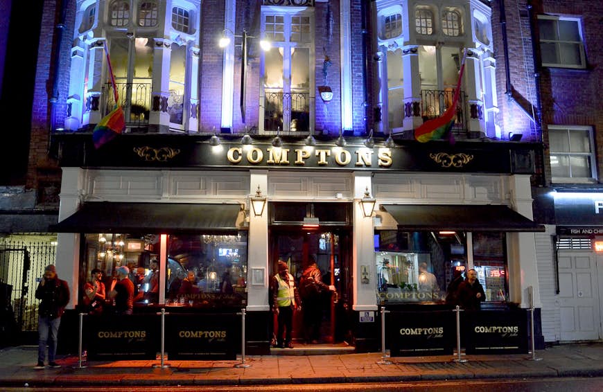 The facade of Comptons of Soho at night; "Comptons" is written in gold letters on a black background; and there are rainbow flags draped outside ornate bay windows on the upstairs floor.