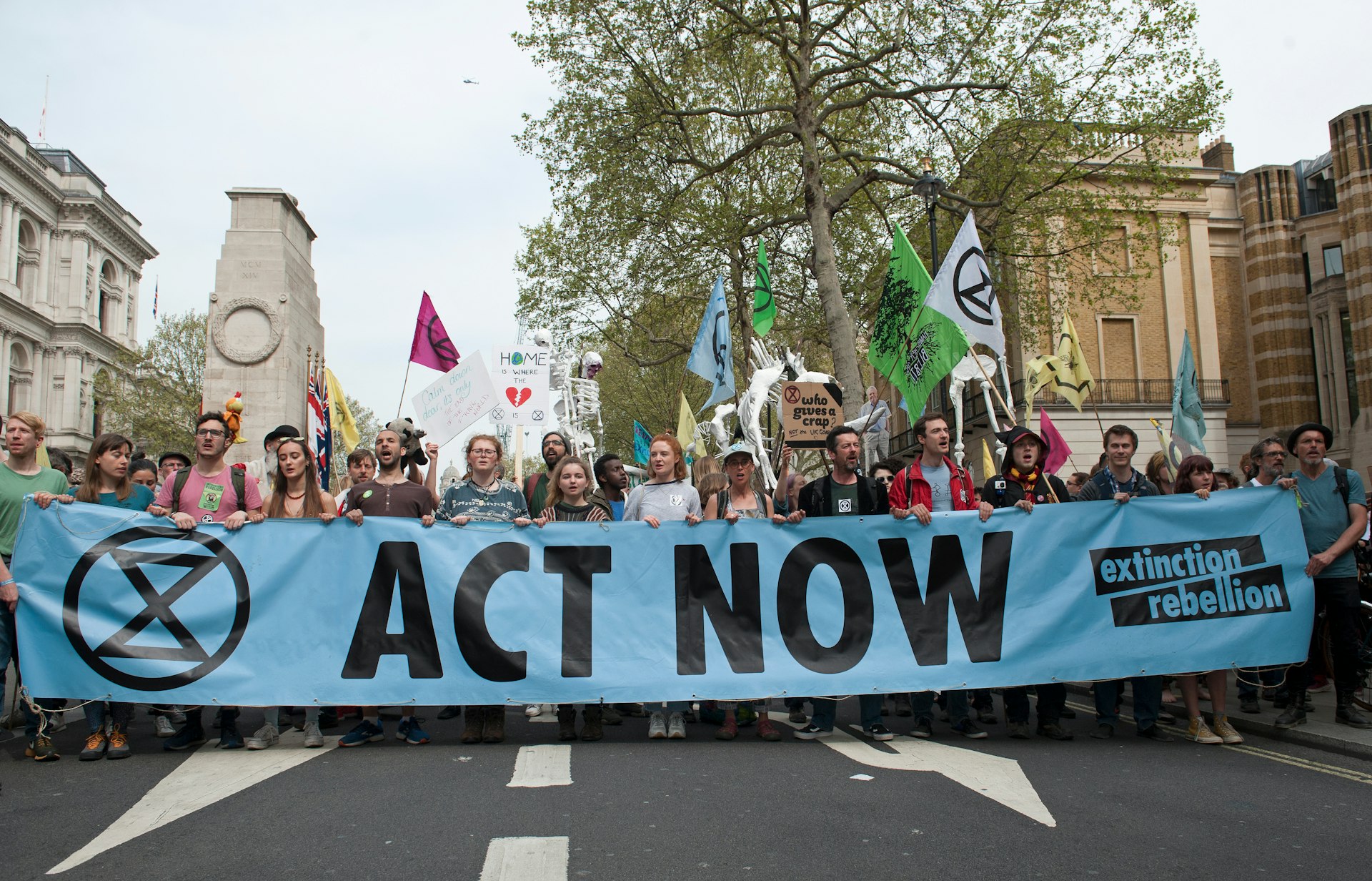 An image of Extinction Rebellion protested in central London. The crowd are carrying a large blue banner reading 'ACT NOW' as well as flags and other smaller signs. 