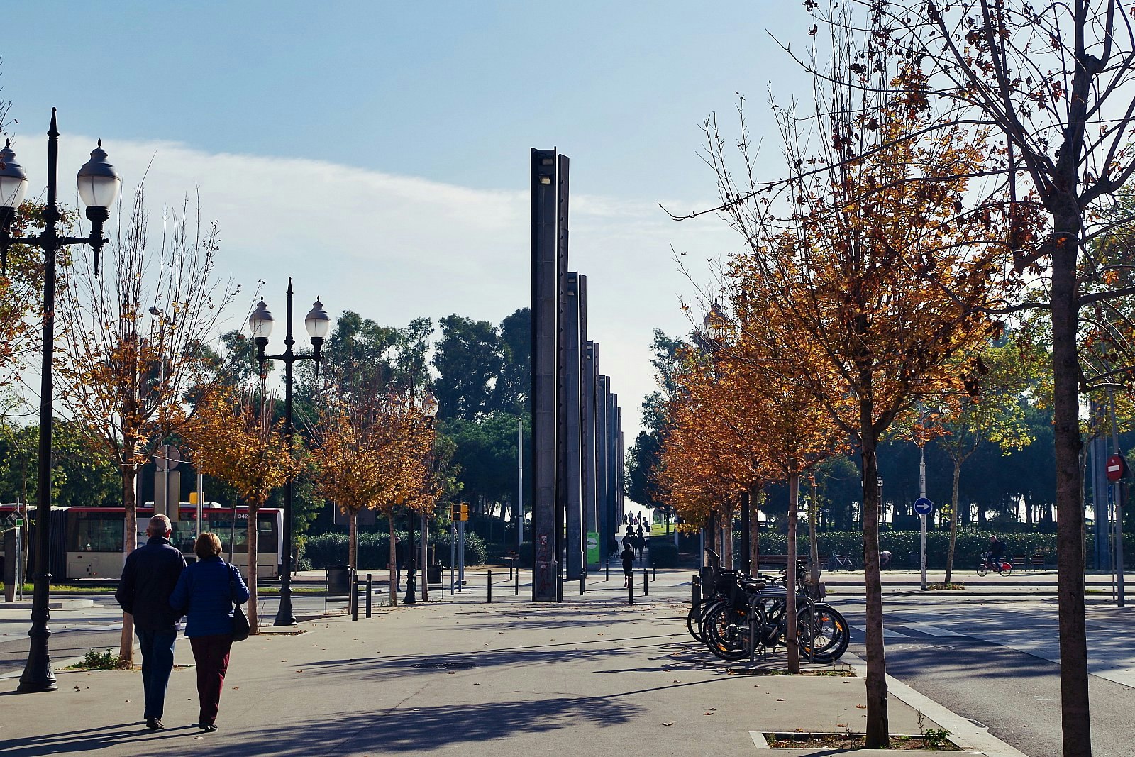 People walking down Rambla del Poblenou; the esplanade is lined with ornate lamp-posts and trees in autumnal colours, and there are sculptural columns further along the Rambla.