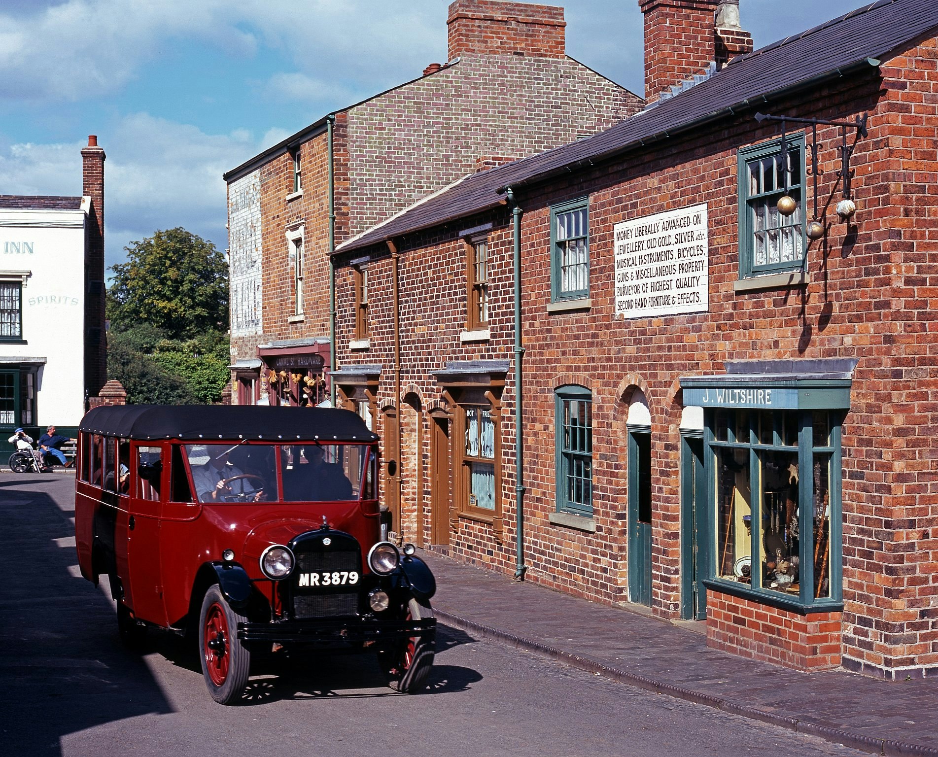 A red vintage car being driven along a recreated historic street of redbrick terraced homes and shops.