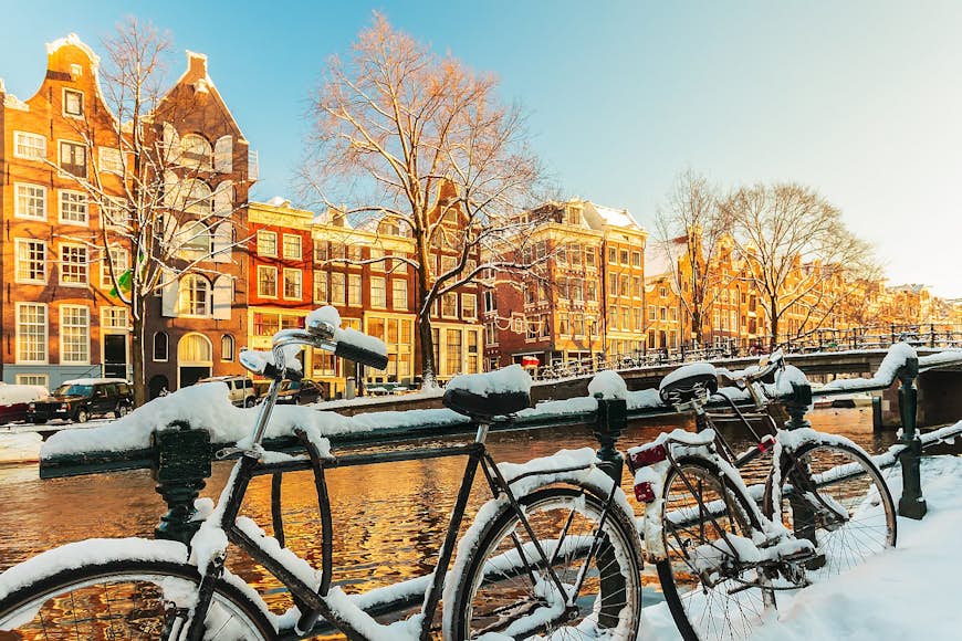 Snötäckta cyklar vilar på ett räcke med en Amsterdam-kanal bakom sig;  på andra sidan kanalen ligger gavelradhus.  Amsterdam vinter