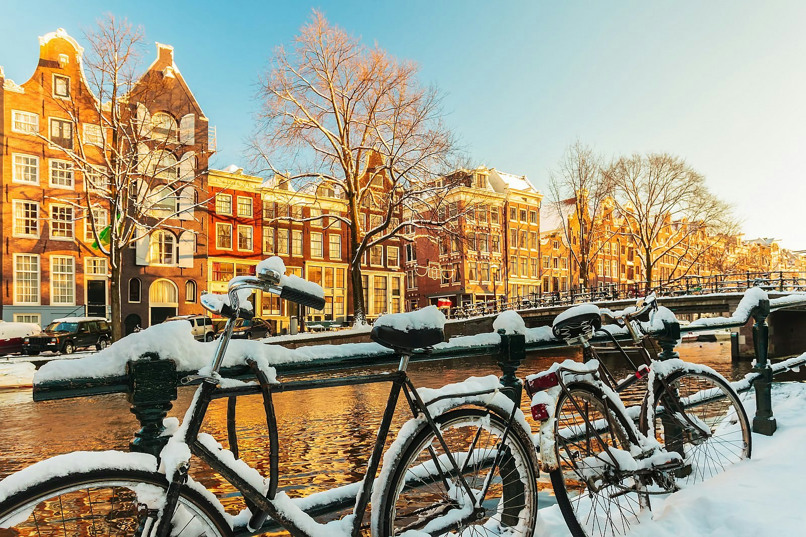Snow-covered bicycles resting on a railing with an Amsterdam canal behind it; on the other side of the canal are gabled townhouses. Amsterdam winter
