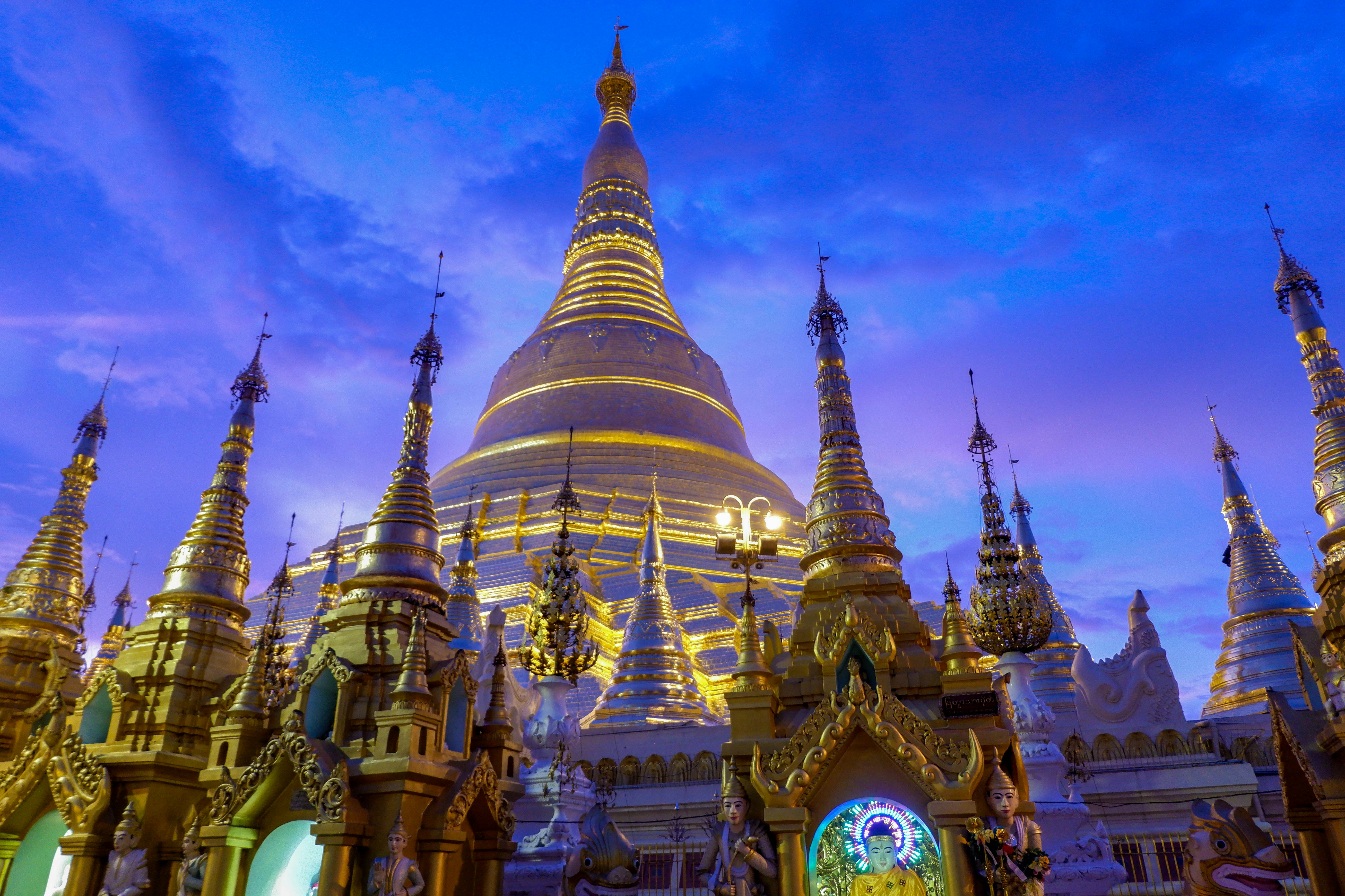 The Shwedagon Pagoda in Yangon at dusk; many small intricately carved, gold-leafed structures surround a huge dome, lit up against the night sky.