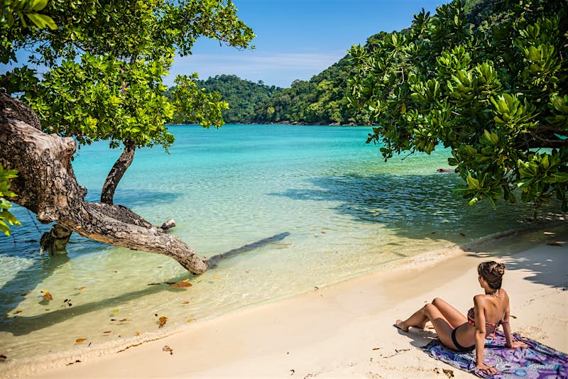 A woman reclines on a tropical beach with vegetation around her and clear blue-green sea lapping on the sand.