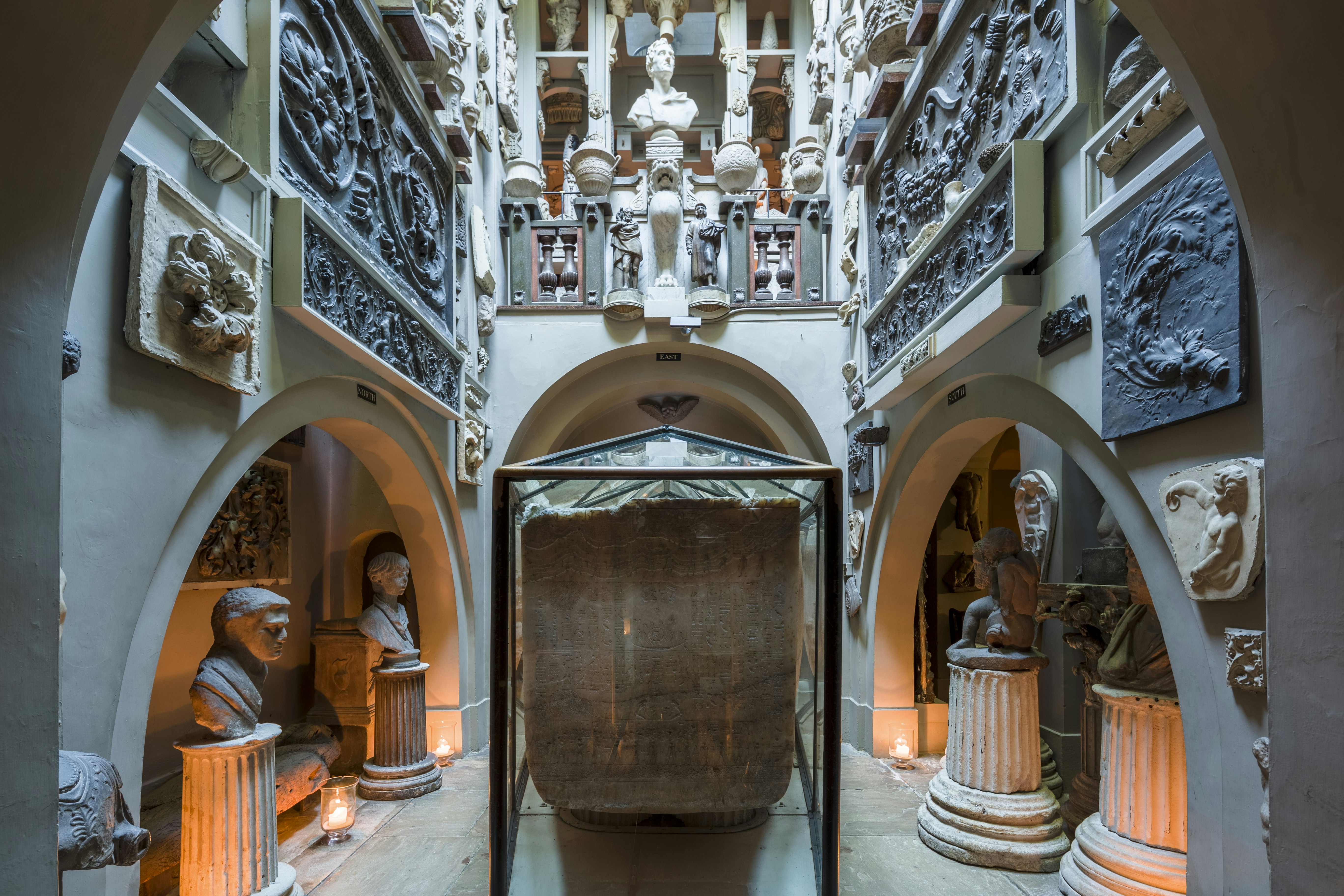 A narrow, high-ceilinged room at Sir John Soane's Museum in London. A sarcophagus in a glass case sits in the middle of the room, which is lined with plaster busts, reliefs and casts.