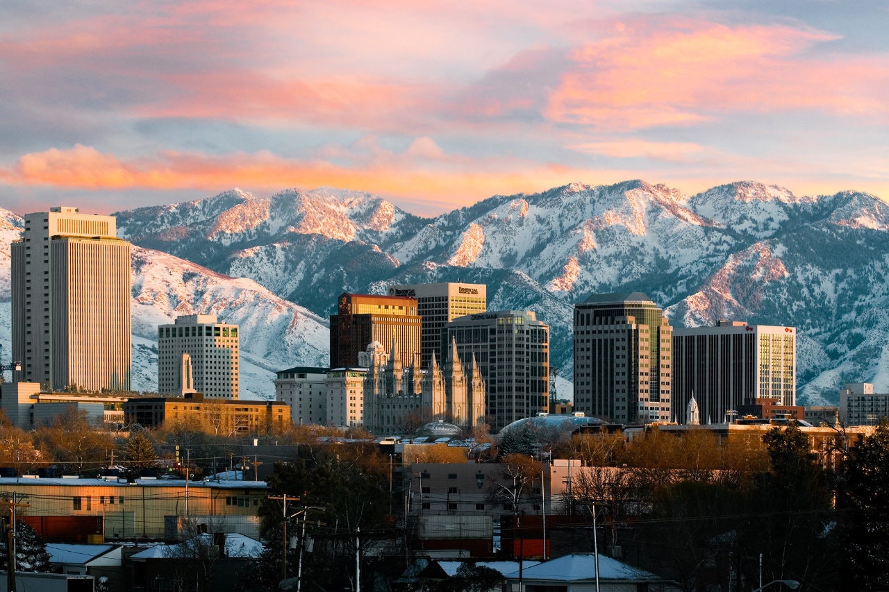 The skyline of Salt Lake City glows white and gold in the early sunset, with the snowy Wasatch mountains close set behind the city and the sky glowing pink overhead