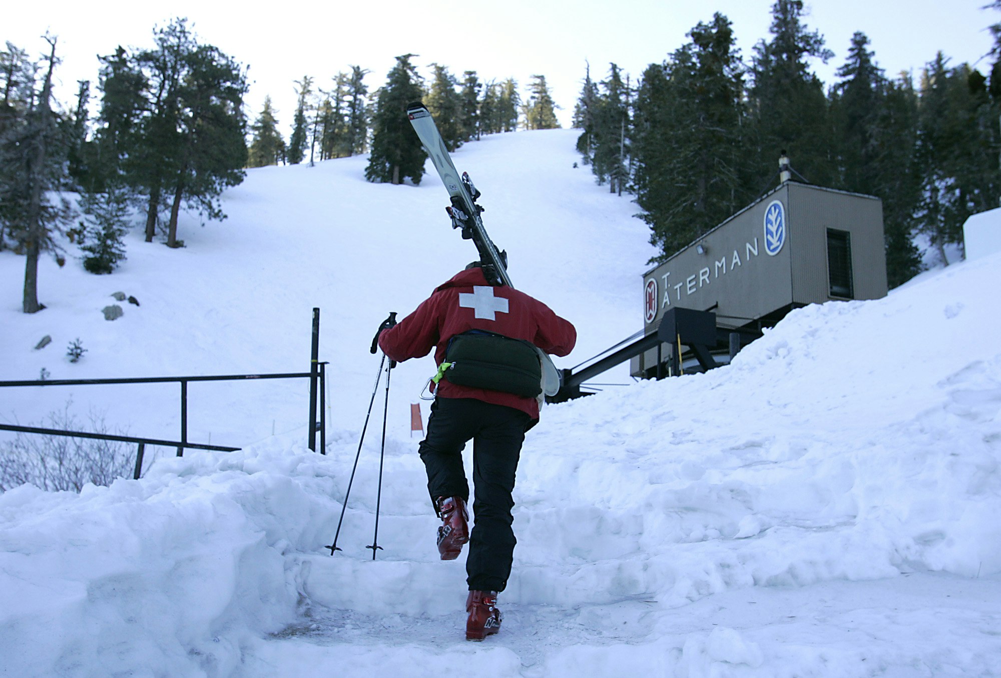 A member of the Mt. Waterman ski patrol in black snow pants, a red ski jacket with a white cross on the back, red boots, and a black fanny pack climbs snowy steps in front of a brown warming hut that reads Aterman in white san serif letters. A snow hill free of tracks rises above the patrolman