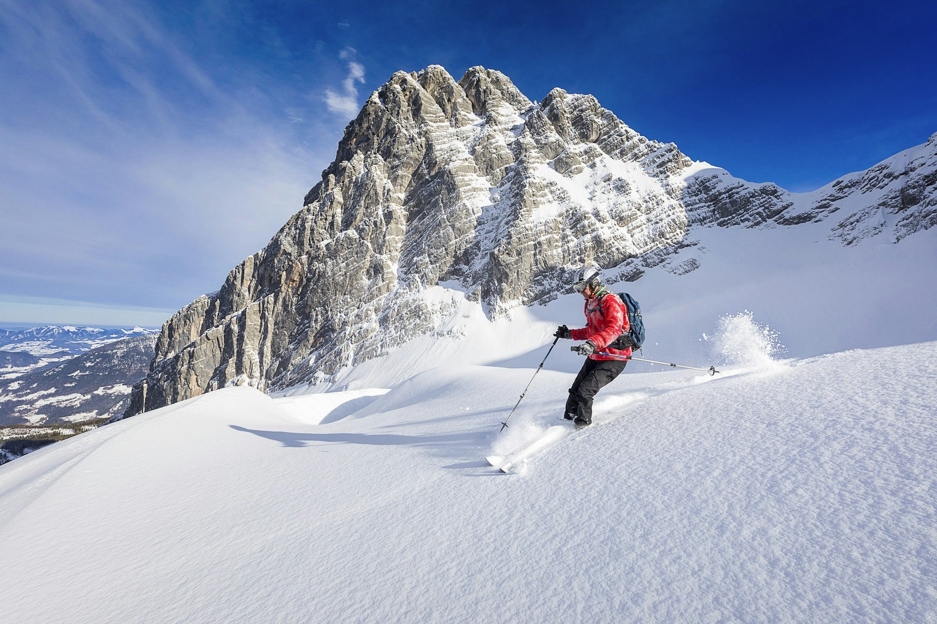 A man sking through powder in the back country of Berchtesgaden National Park; he's in a large track of virgin powder with a large pyramid rocky summit rising in the background.
