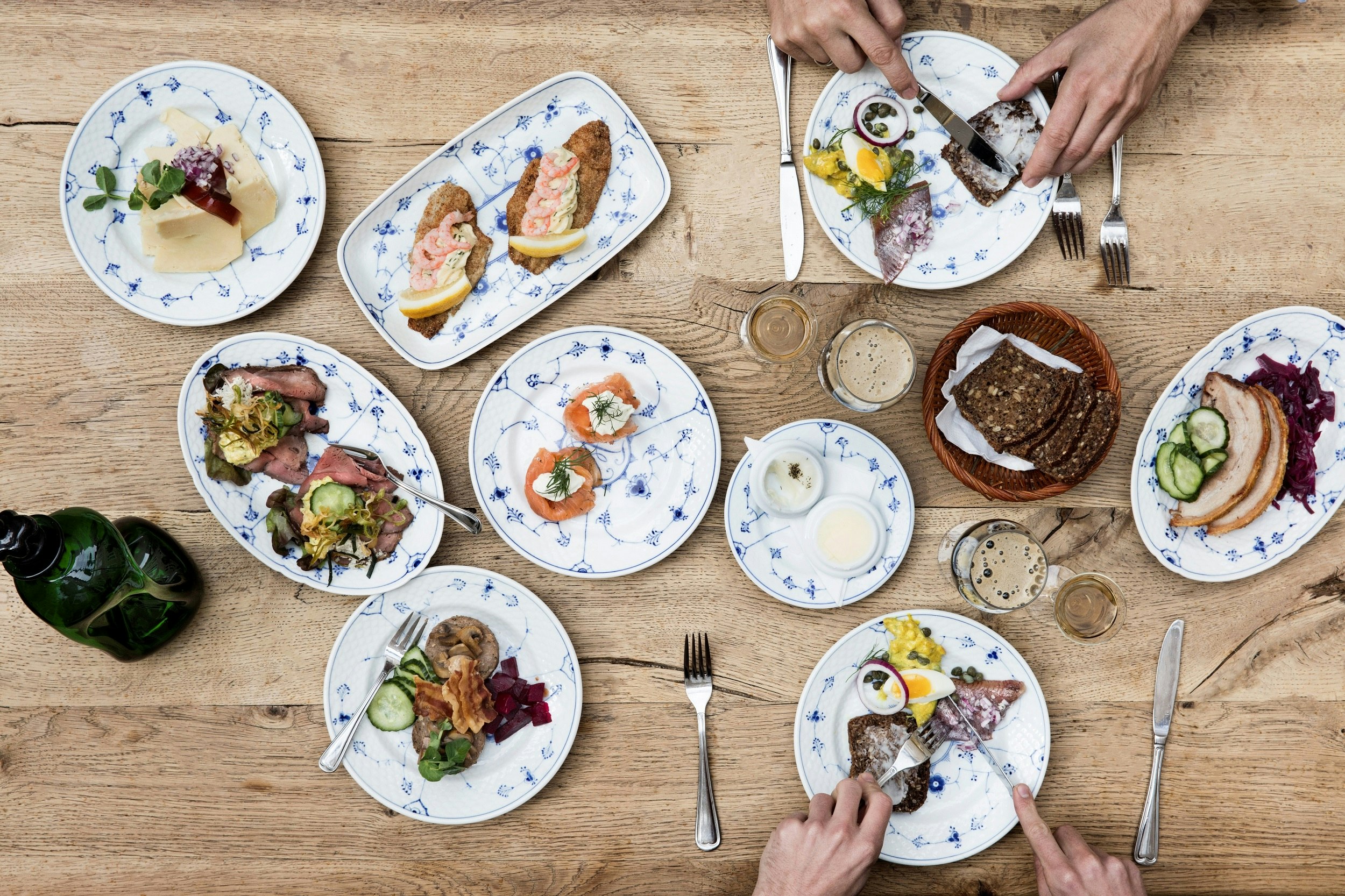A warm oak table is laden with plates of smørrebrød, with two people's hands visible on each side; both people are using knives and forks to eat.