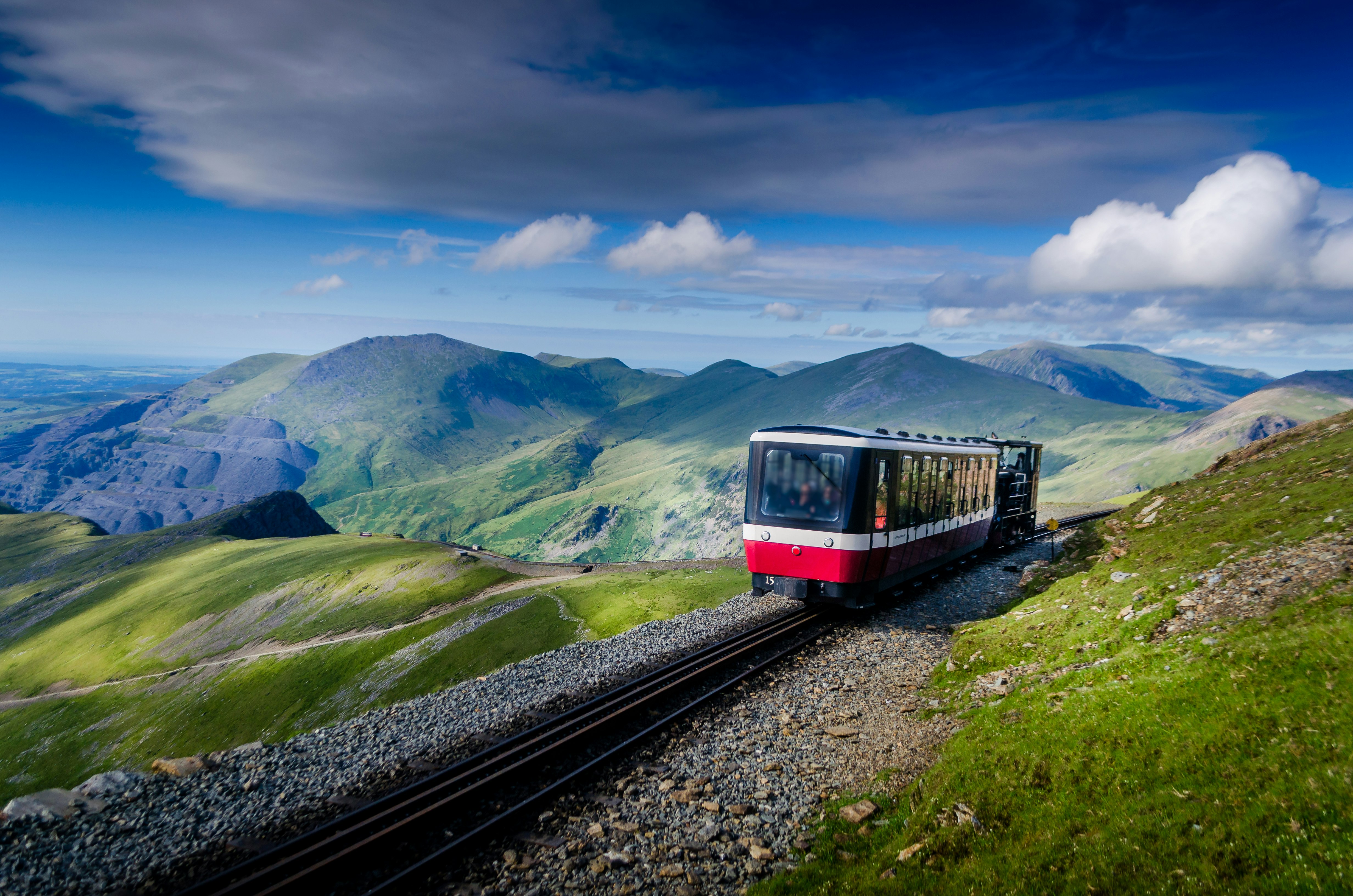 A small red, narrow-guage tourist train rides the rails towards Snowdon in Snowdonia National Park, as sun bathes the green peaks of the range.