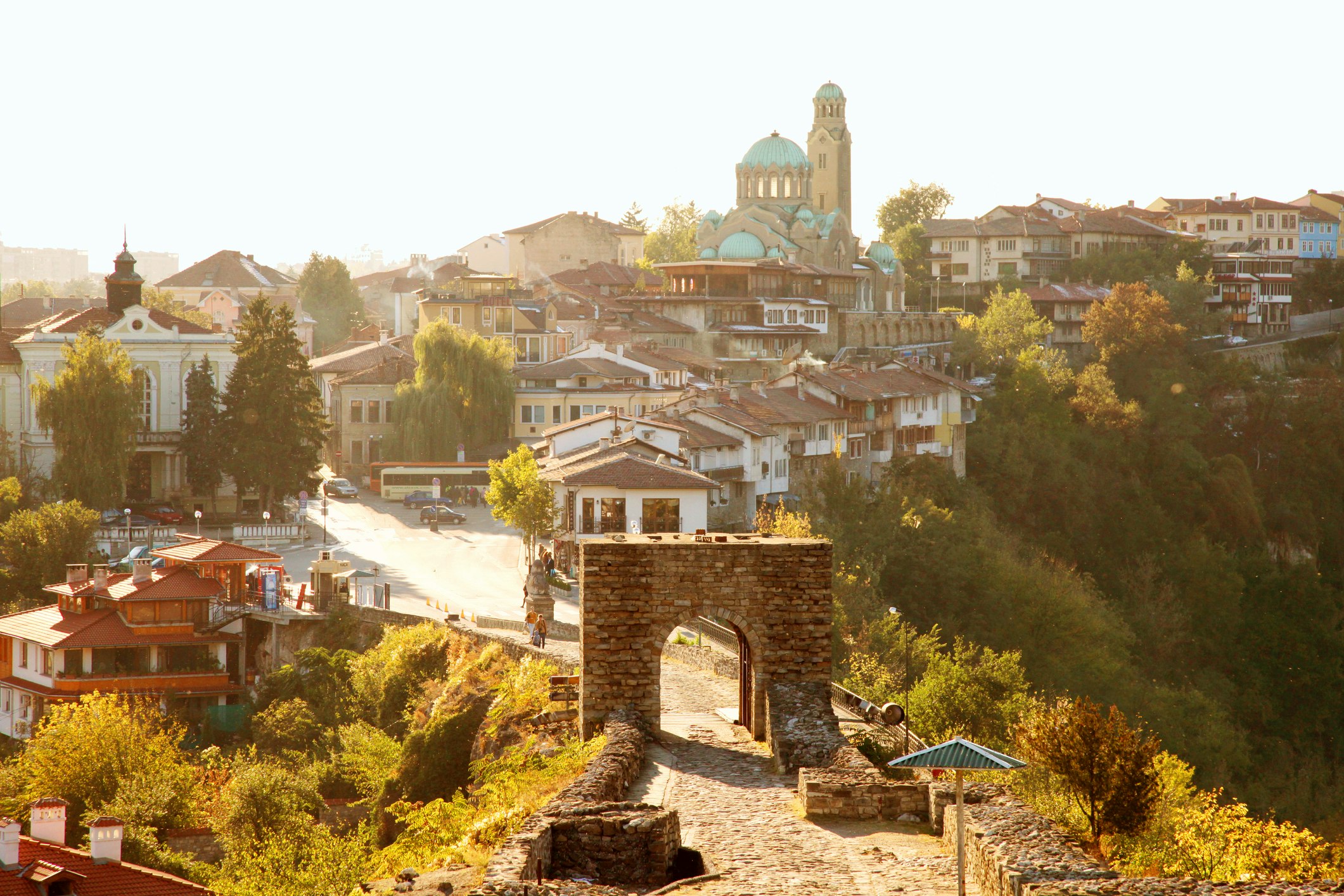 A warm, slightly hazy shot with the tones of a vintage photo showing the hillside town of Sofia, Bulgaria. A simple stone arch in the foreground guides the viewer's eye up a broad street between the medieval houses and towards the large Saint Aleksandar Nevski Cathedral on the hill in the central background.