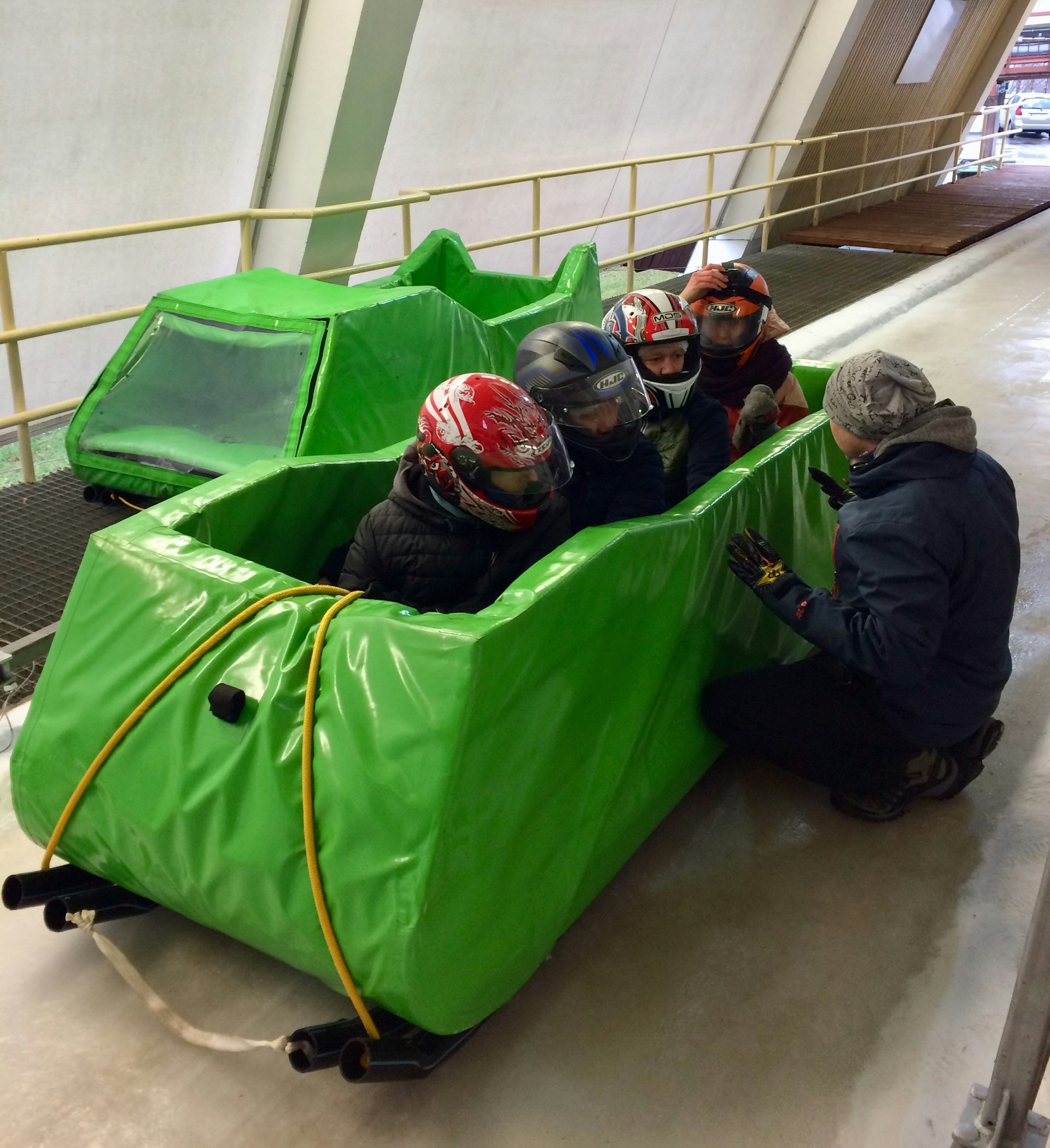 Four people wearing helmets sit in a toboggan-like vehicle which is covered in thick green padding. An instructor kneels nearby