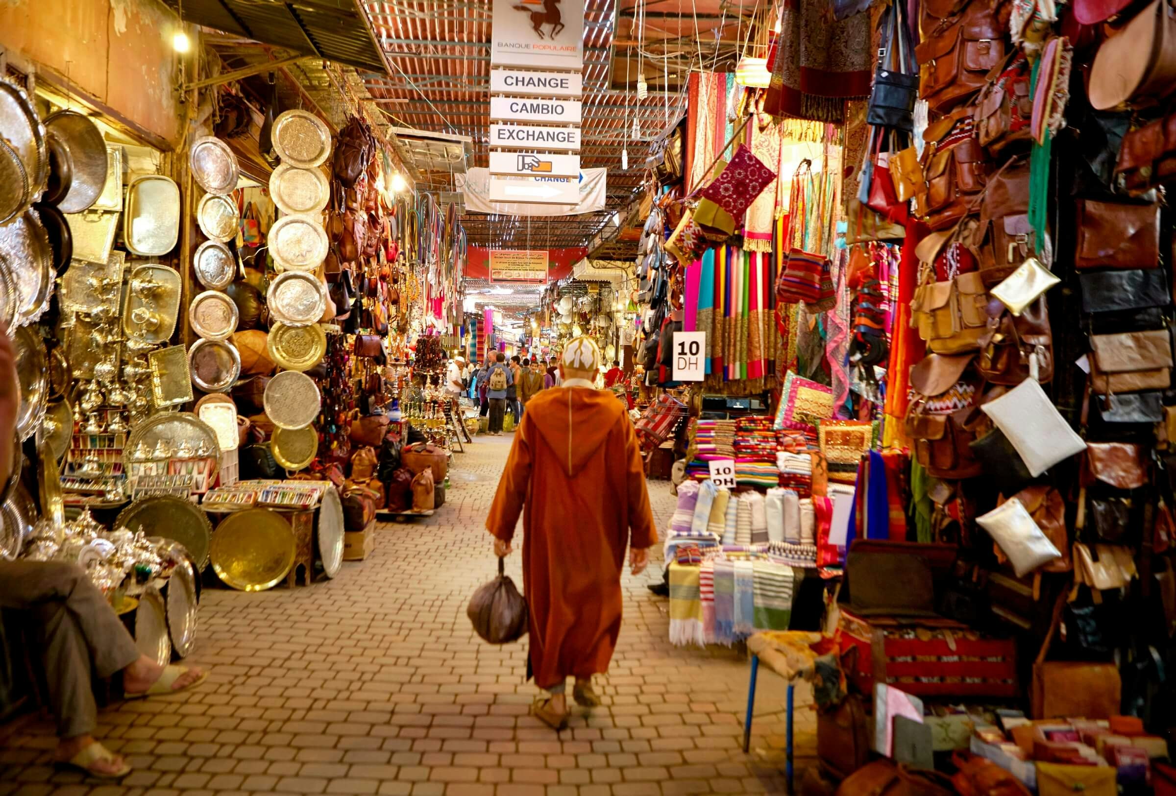 A man in traditional Moroccan robe called a Djellabah walks away from the camera. Stalls selling plates, bags and scarves line either side.