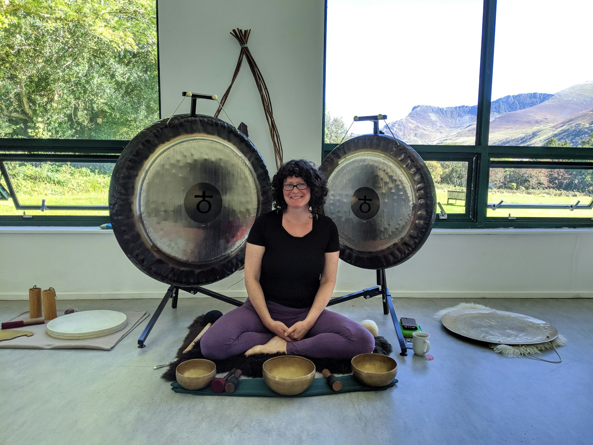 A woman sits cross legged in front of large gongs and has metal bowls at her feet.