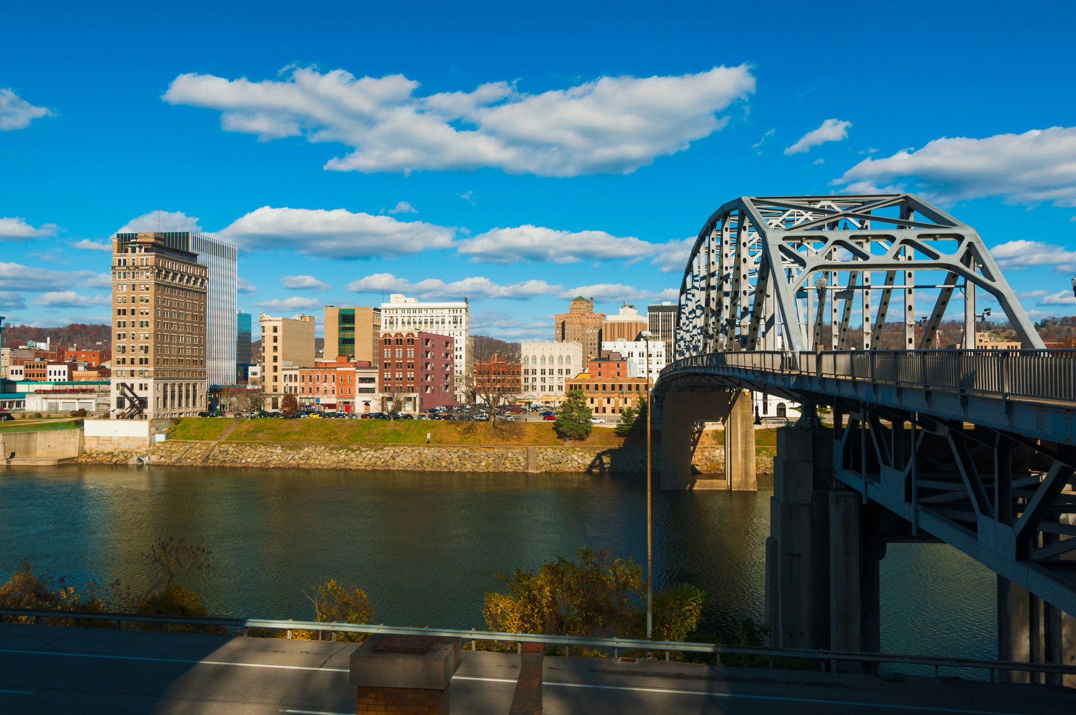 A view of the Charleston, West Virginia Skyline from across the Kanawha River. On the right side of the frame is the South Side Bridge, extending from the Amtrak station to the city center. The skyline is mostly made up of low, multi-story brick and masonry early skyscrapers in beige or neutral tones. The river is a deep greyish blue. The sky is bright and full of fluffy white clouds. 