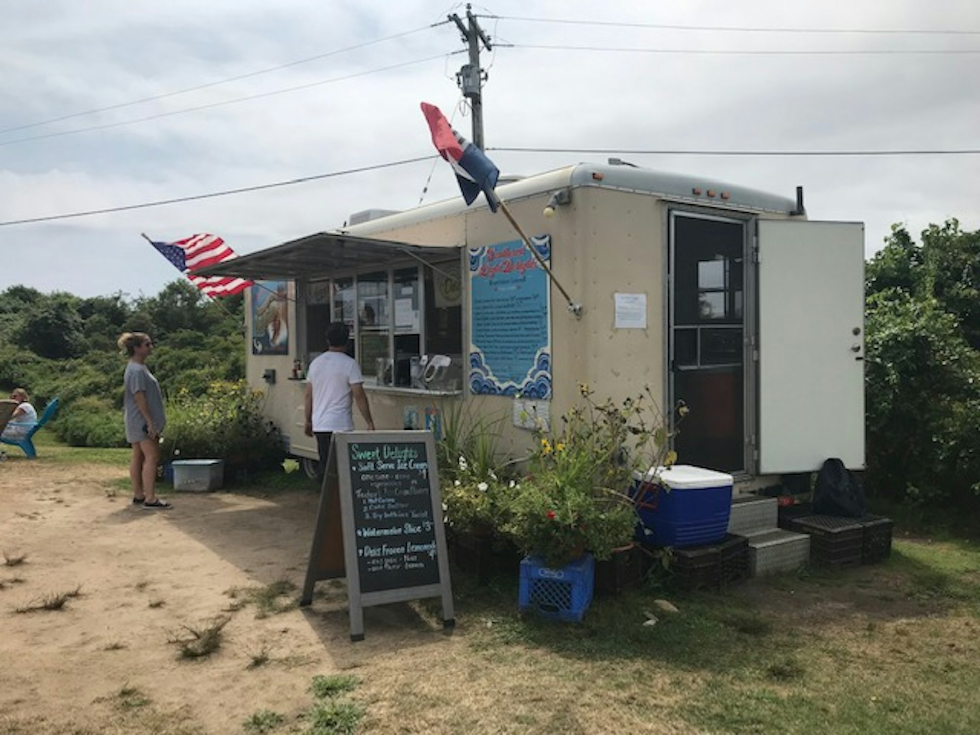 A food stall called 'Southeast Light Delights'. There's a man in a white t-shirt ordering food, and an American flag draped from the stall is blowing in the wind. 