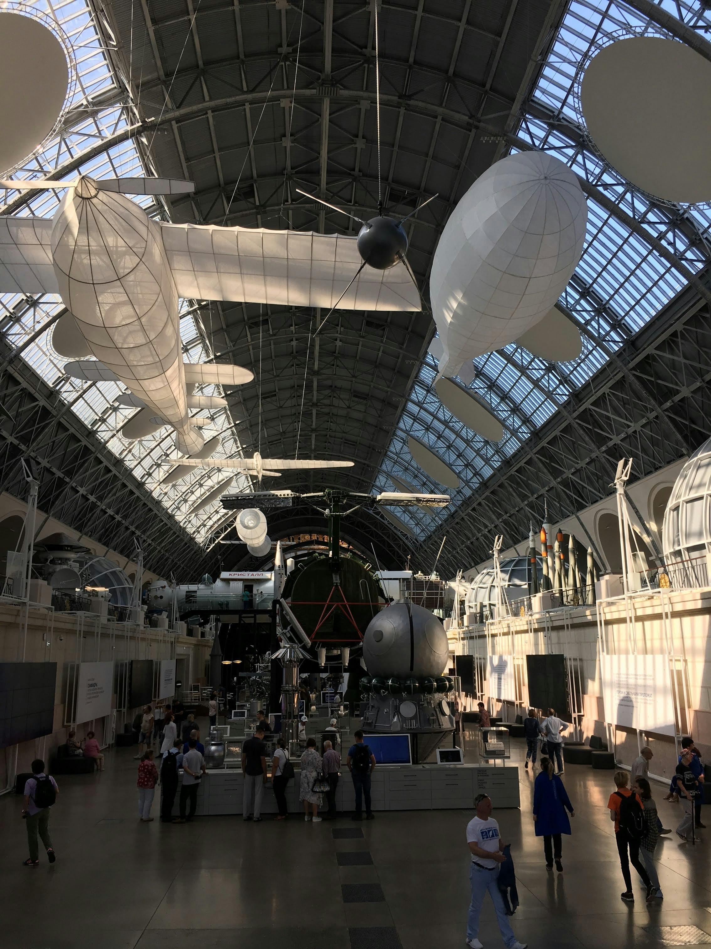 A large room with a steel and glass roof. There are objects hanging from the ceiling including a plane and a blimp, which both appear to be made of paper