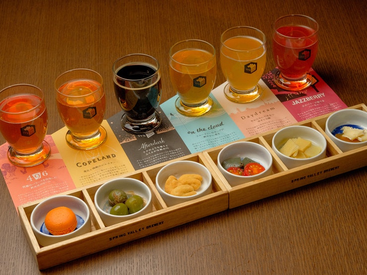 A row of six beers ranging in color from pinkish red to dark black to golden yellow are lined up on colorful descriptors, each with a small white circular ceramic dish in front of it containing a small snack selected for flavor pairings, like olives, a macaron, or tomatoes