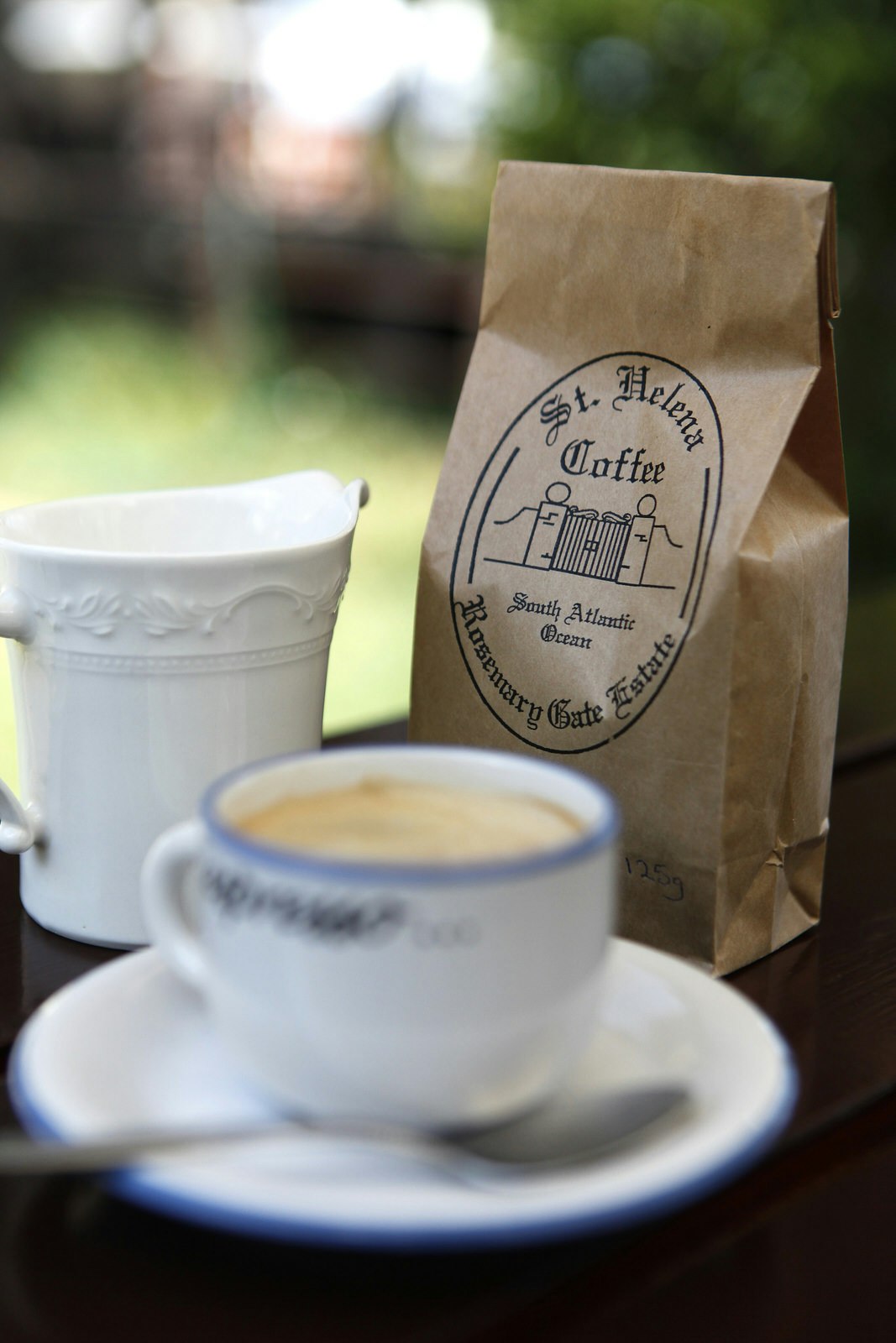 A blurred white cup with a blue rim - full of coffee - sits on a white saucer with spoon next to a white china cream jug; behind the pair (in focus) is a brown paper bag with a logo reading St Helena Coffee.