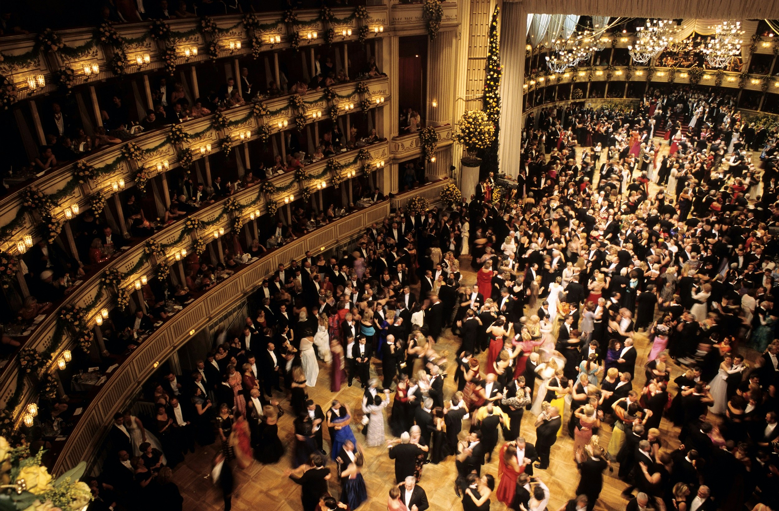 A shot downwards onto a busy ballroom dance floor, packed with couples dancing together. Men are wearing dark suits; women are in evening dresses of many different colours. Balcony boxes are full of people, and chandeliers hang from the ceiling