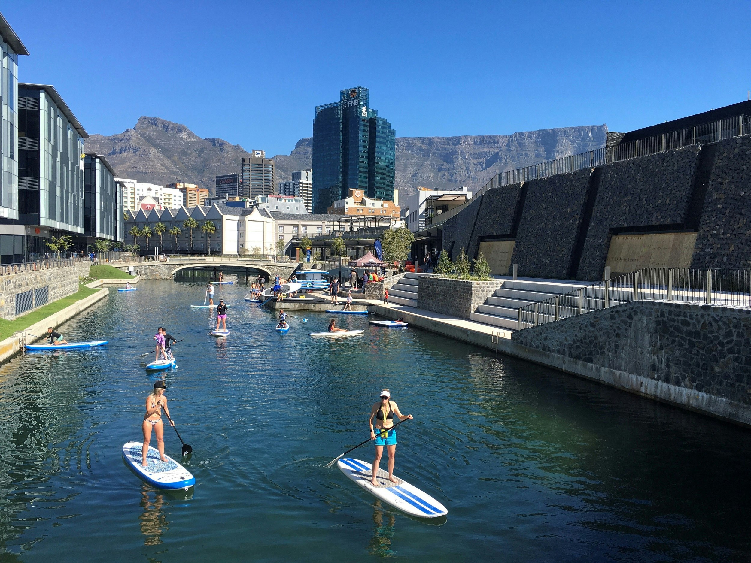 A dozen paddle boarders cruise along a narrow section of the V&A Waterfront; in the background is Table Mountain.