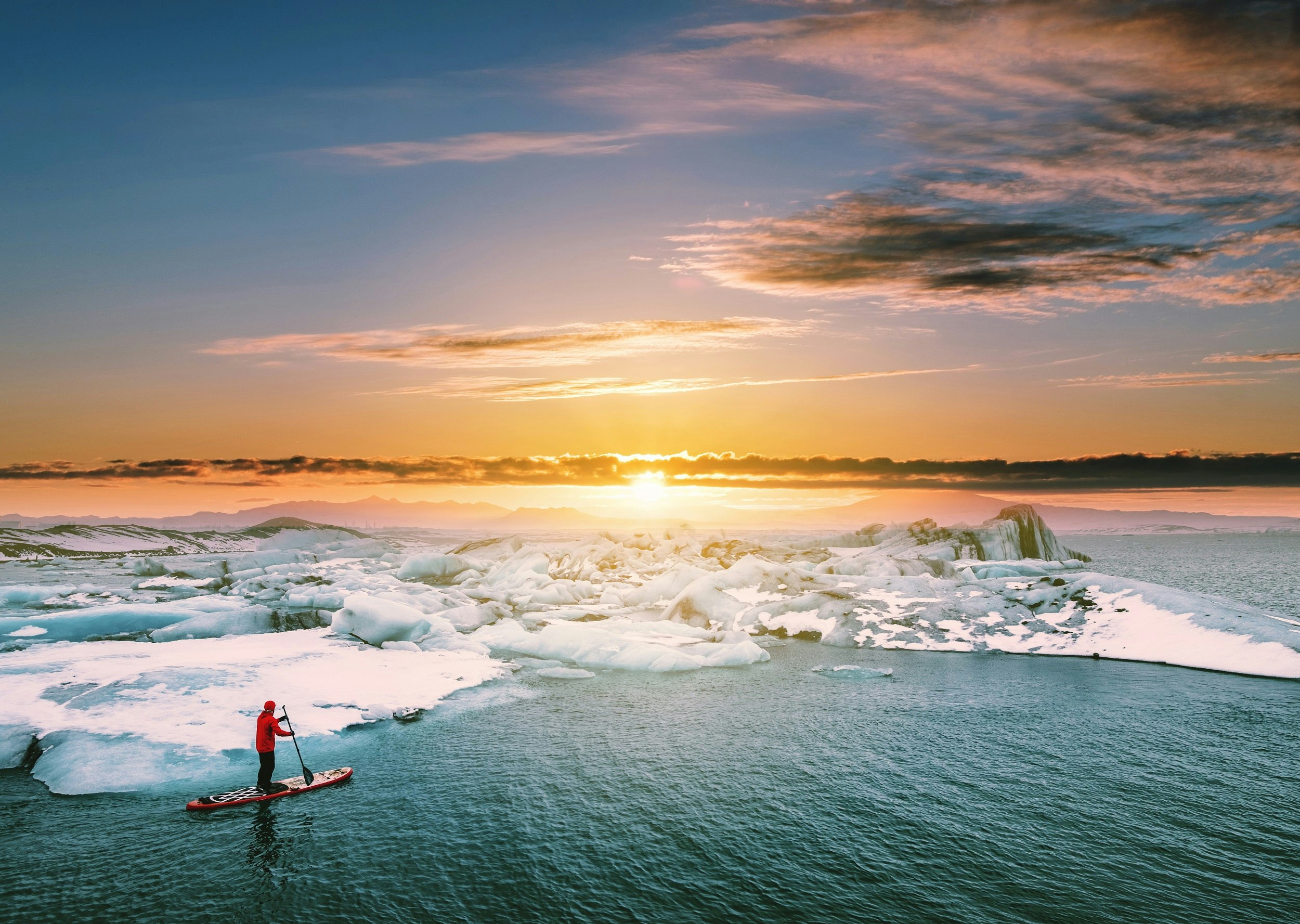 A lone stand-up paddleboarder in survival gear makes their way along the edge of a glacier in the sea, with the sun setting behind some clouds in the distance.