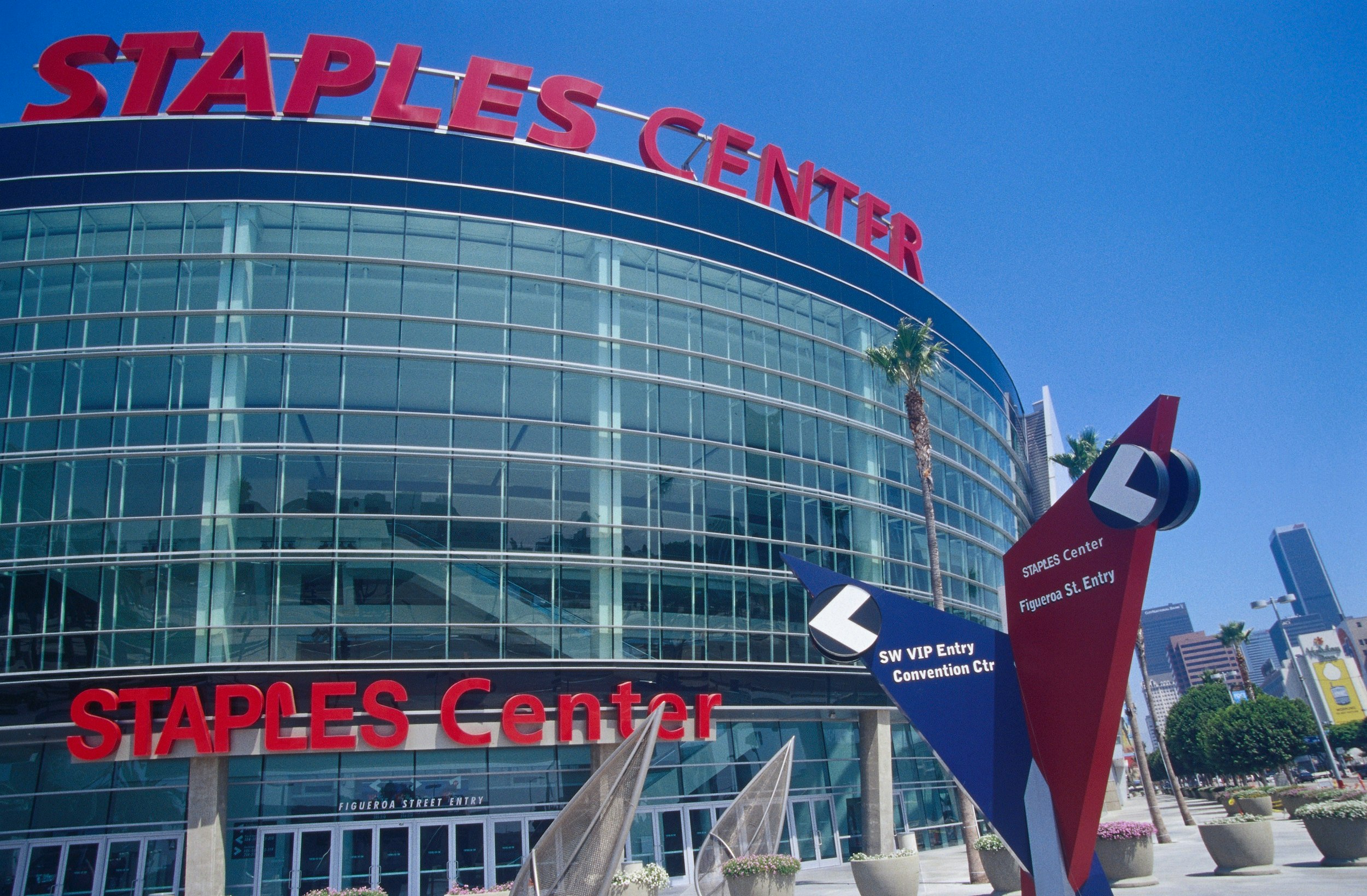 An exterior shot of Staples Center sports arena; the massive, curved glass structure has large red letters atop, and above the entrance, which state its name.