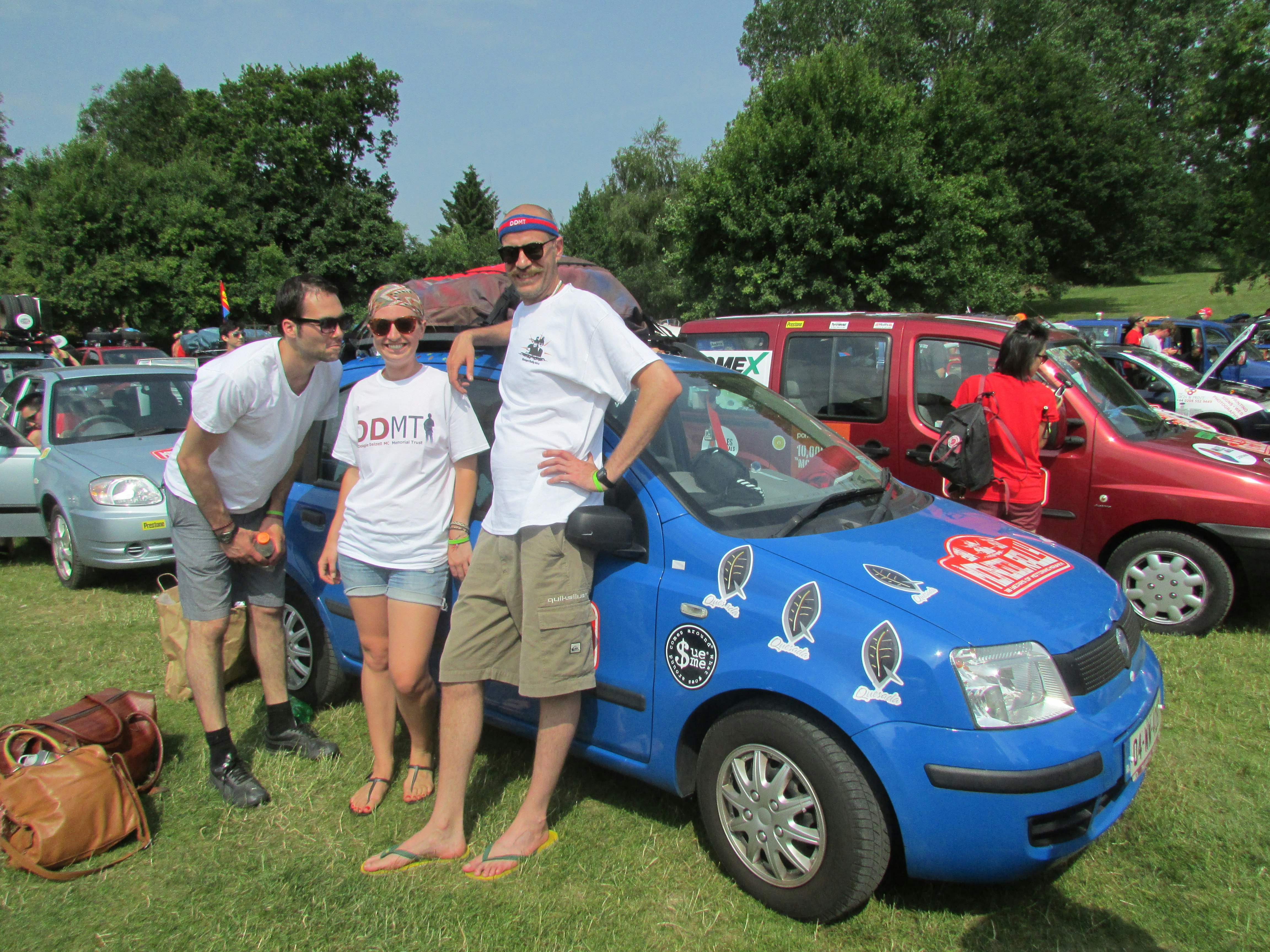 Two men and a woman in matching white Mongol Rally t-shirts and black wayfarer sunglasses stand in front of a blue European minivan covered in stickers at the starting line of the race