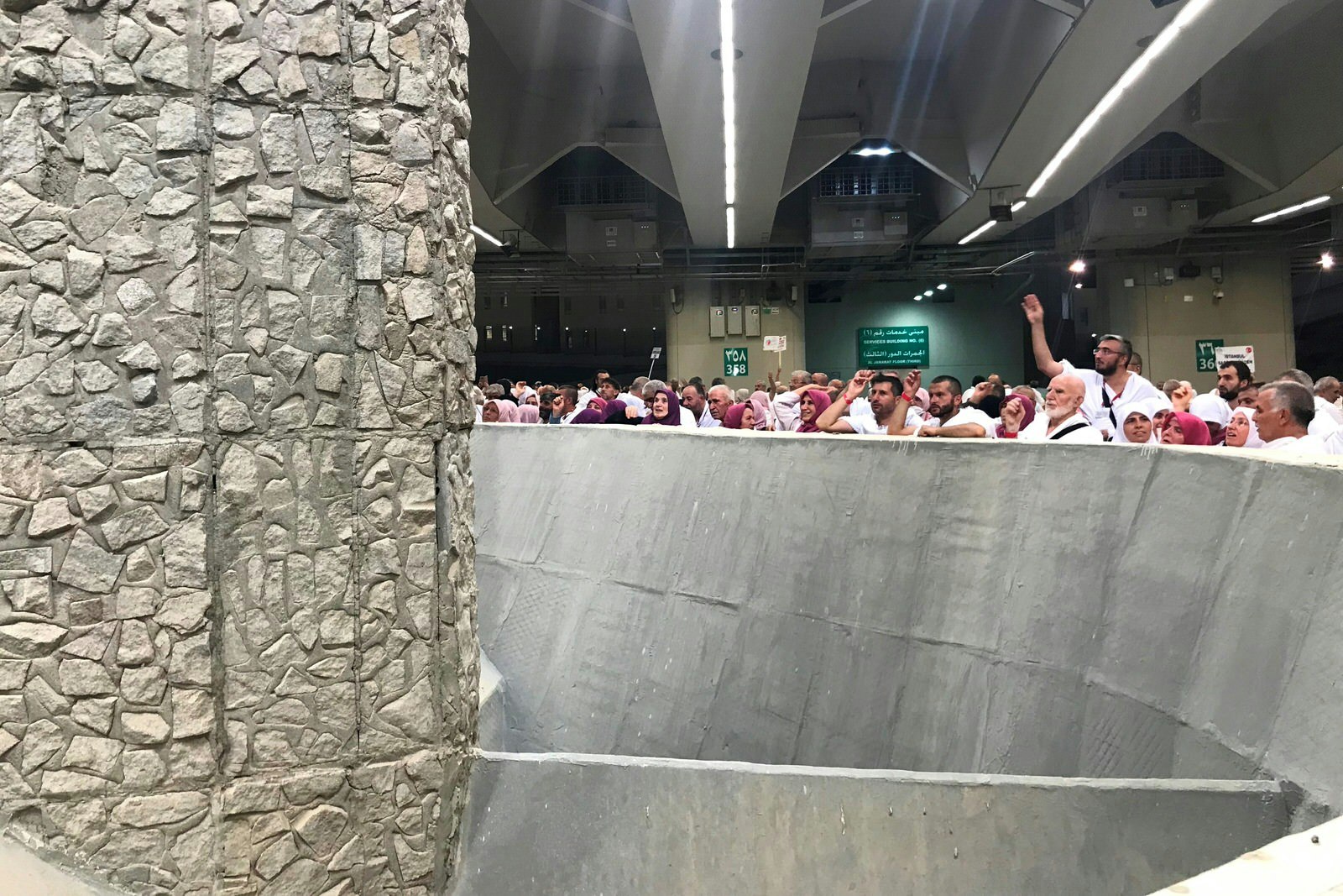 A large pill constructed from large angular rocks in a cement matrix sits surrounded by a concrete wall, behind which Muslim pilgrims throw stones.