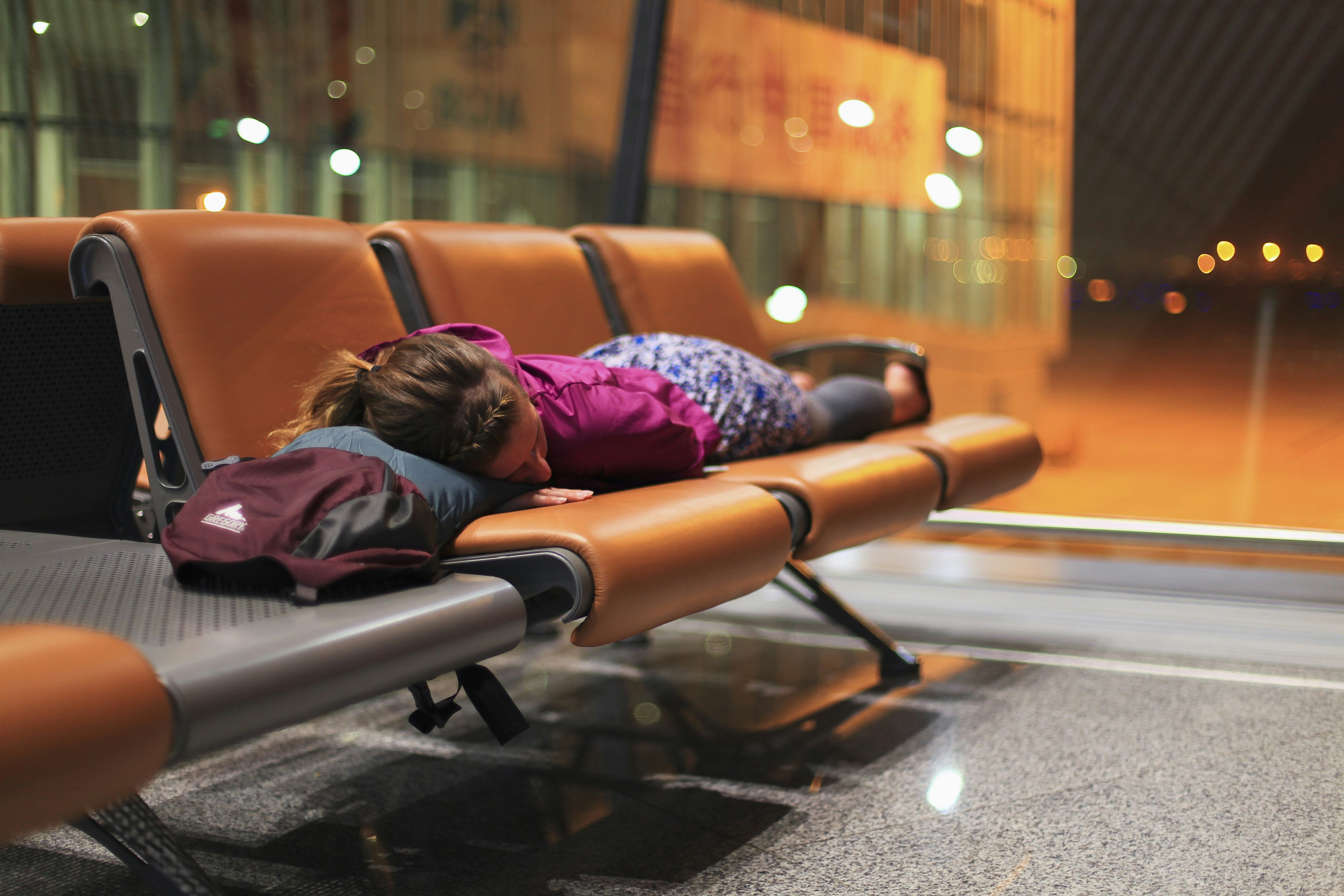 A woman sleeps on an orange bench in an airport in front of a large window. 